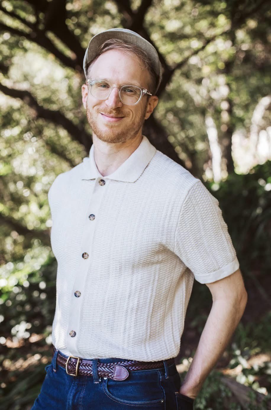Smiling man with glasses, wearing a light-colored shirt, hat, and patterned belt, standing outdoors with trees in the background.