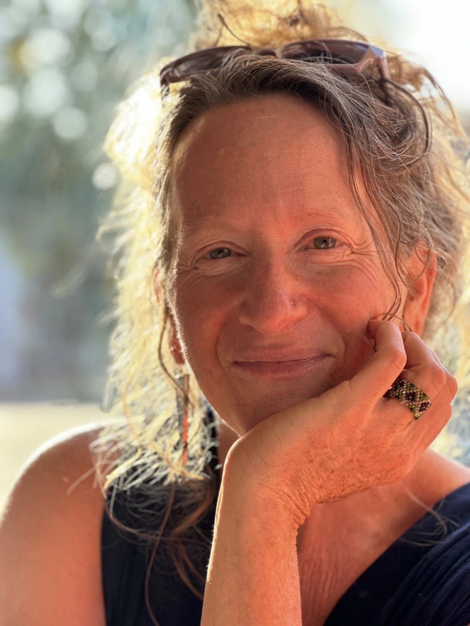 A woman with wavy hair wearing sunglasses on her head, resting her chin on her hand and smiling in natural light.