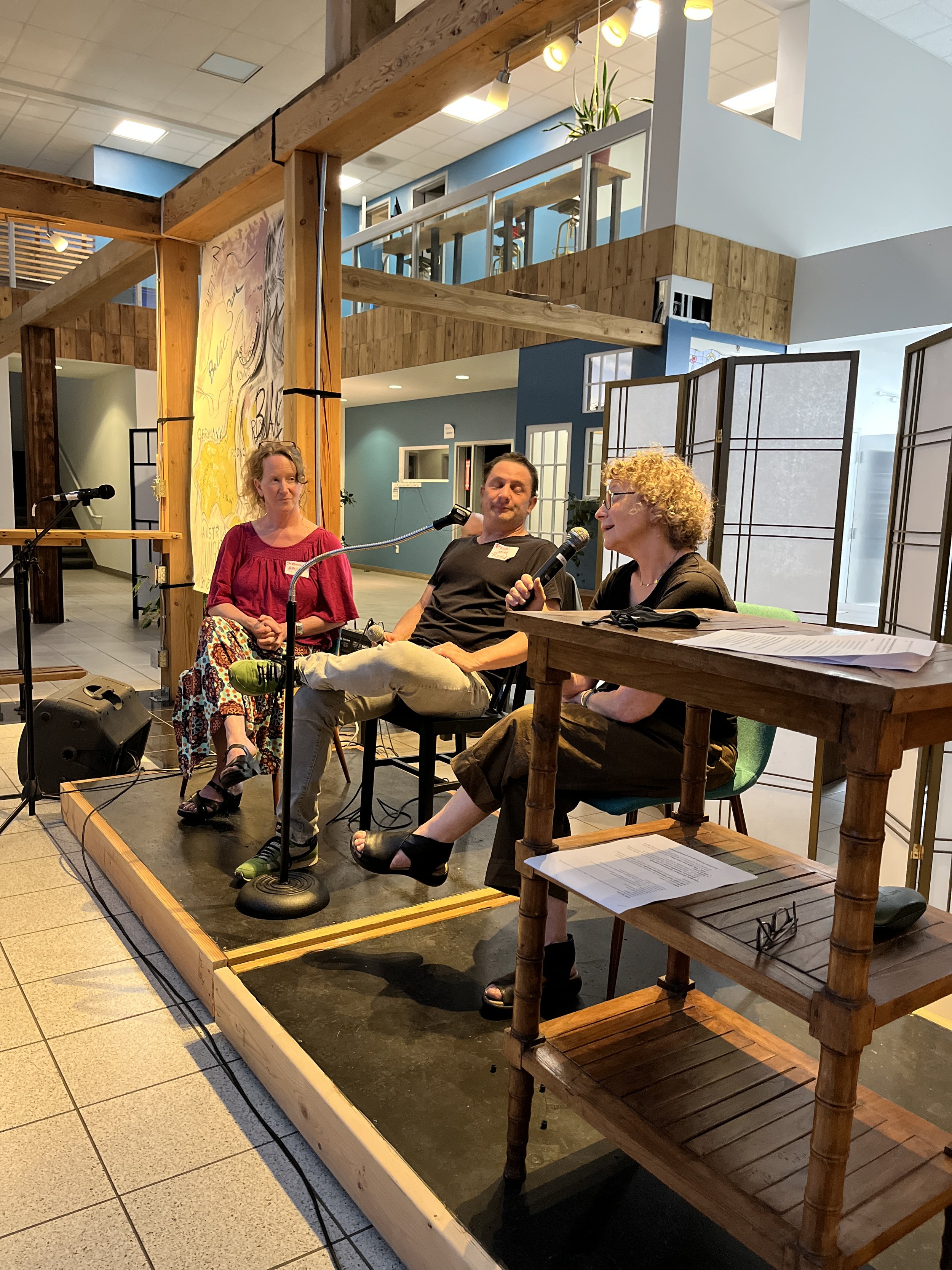 Three people sitting on a small stage, engaged in a panel discussion or interview, with microphones in front of them, in a room with wooden accents and a modern, open design.