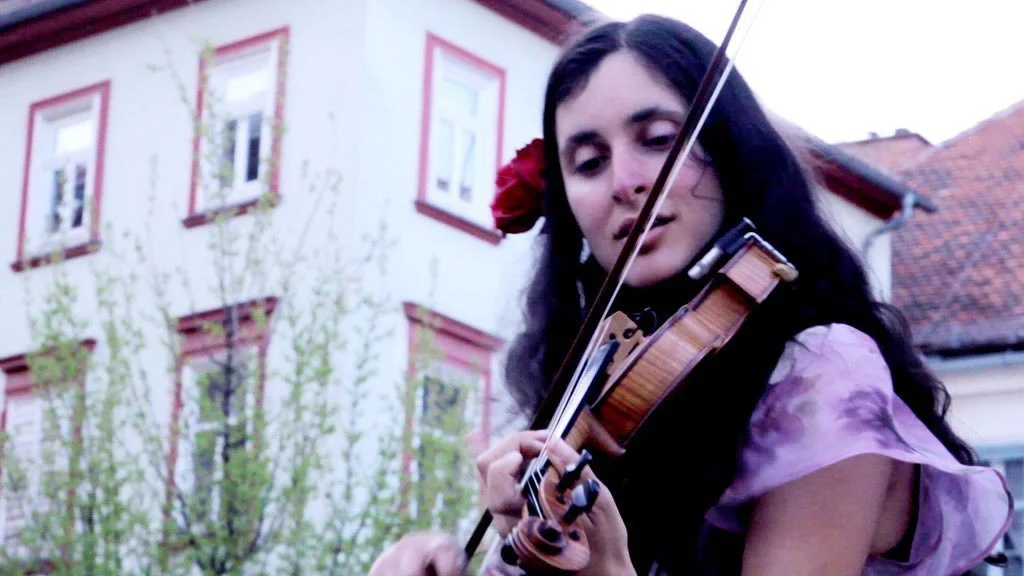 A woman with long dark hair, wearing a pink blouse and a red flower in her hair, playing a violin outdoors in front of a building with white walls and red window frames.