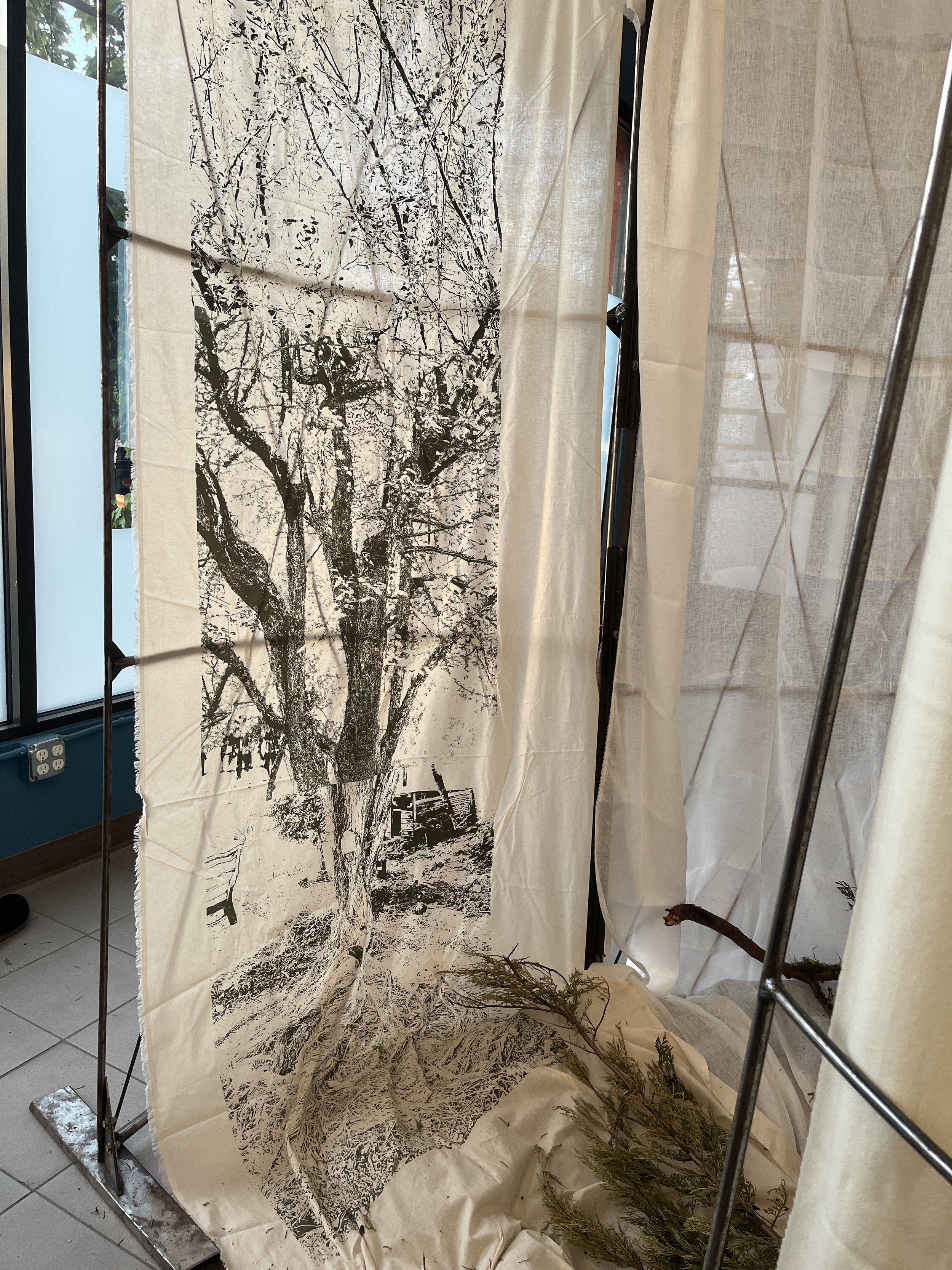 A large black and white print of a tree on fabric, displayed on a metal frame indoors, with some branches and leaves on the floor.