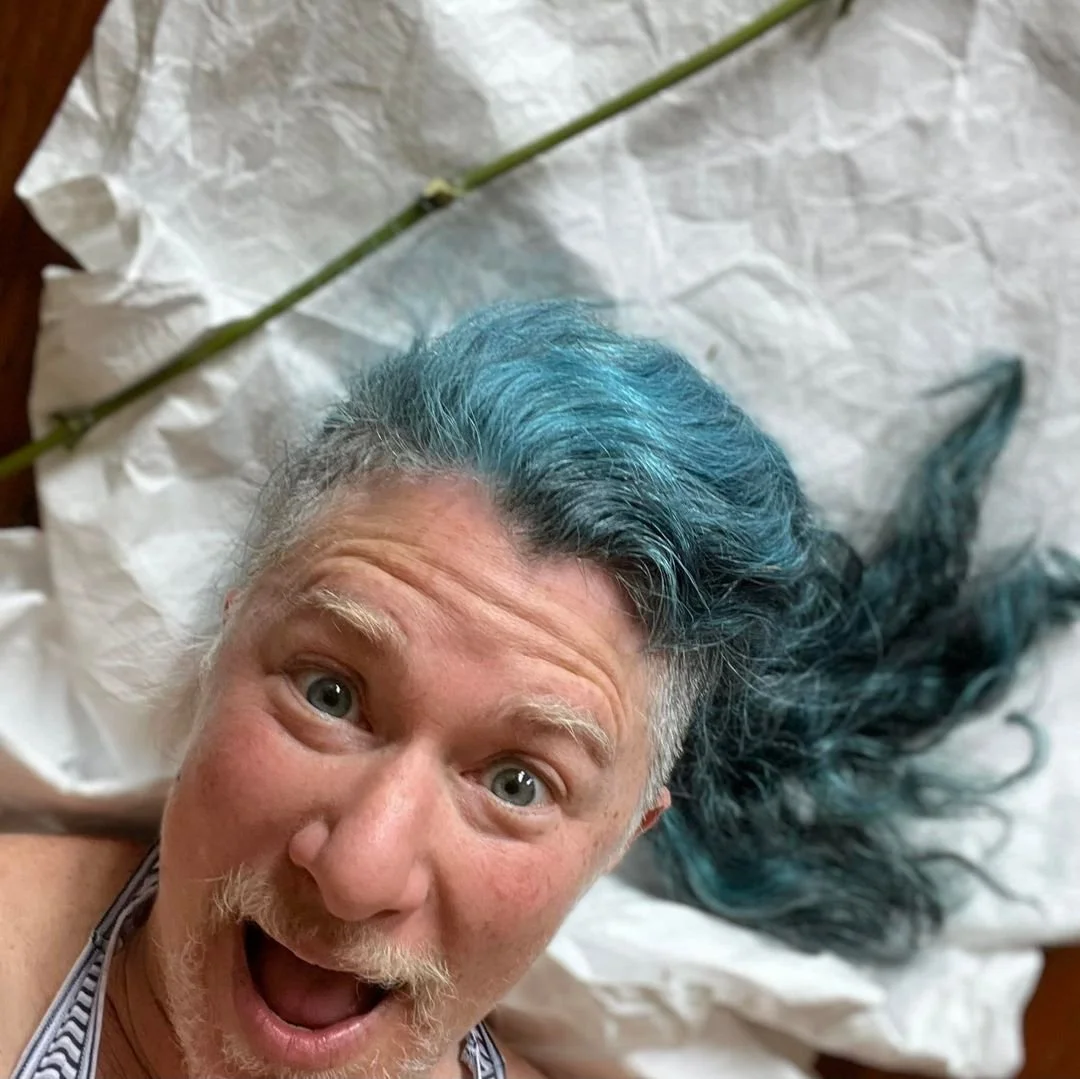 A man with gray hair dyed blue on top, lying on crumpled white paper, taking a selfie with a surprised and happy expression.