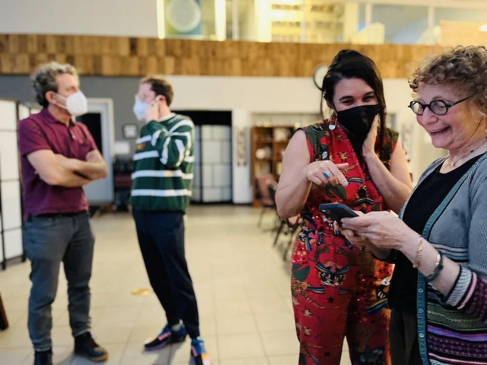 Four people wearing masks. Two men on the left are talking, one in a maroon shirt and gray pants, the other in a green and white striped shirt. On the right, two women are looking at a phone, one in a colorful dress with dark hair and a black mask, t