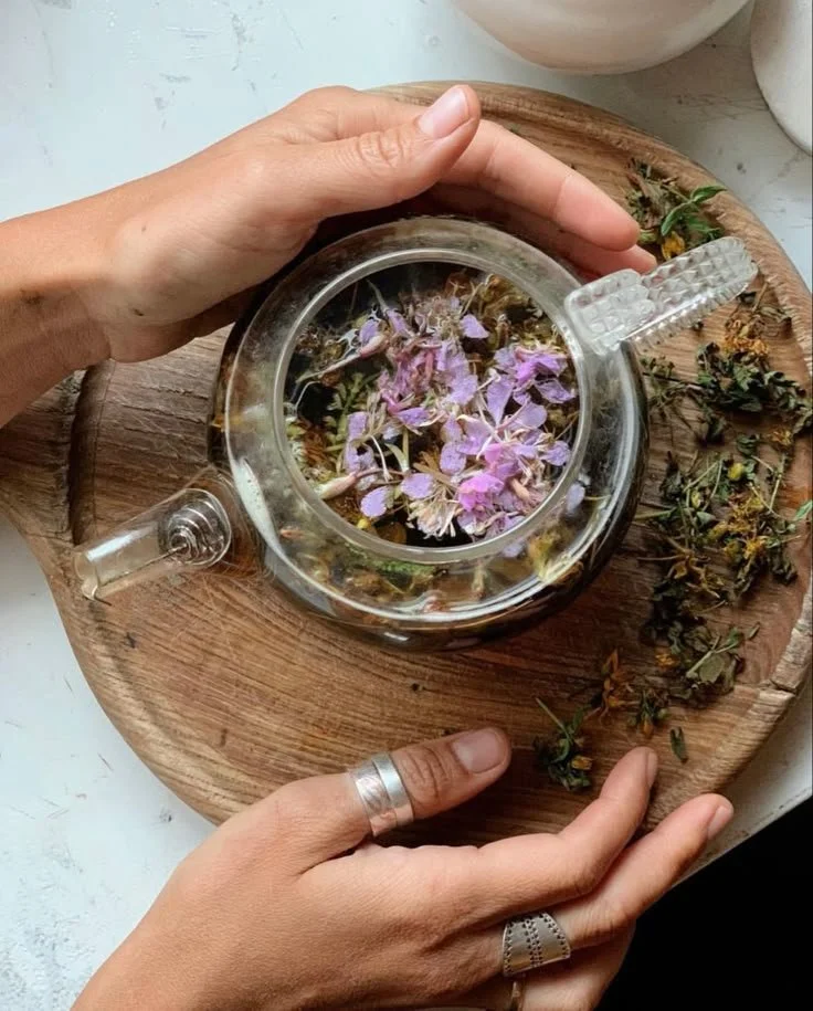 A person's hands with rings hold a clear glass teapot containing blooming tea with purple flower petals, placed on a round wooden tray with dried herbs around it.
