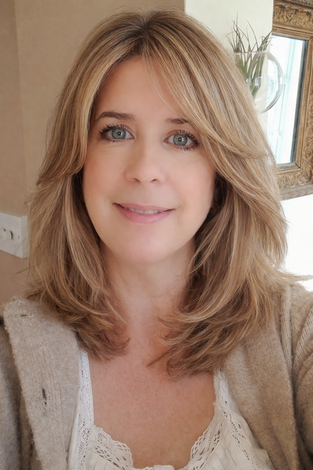 A woman with shoulder-length blonde hair and blue eyes smiling at the camera, wearing a beige cardigan and a white lace top, with a window, wall, and decorative mirror in the background.