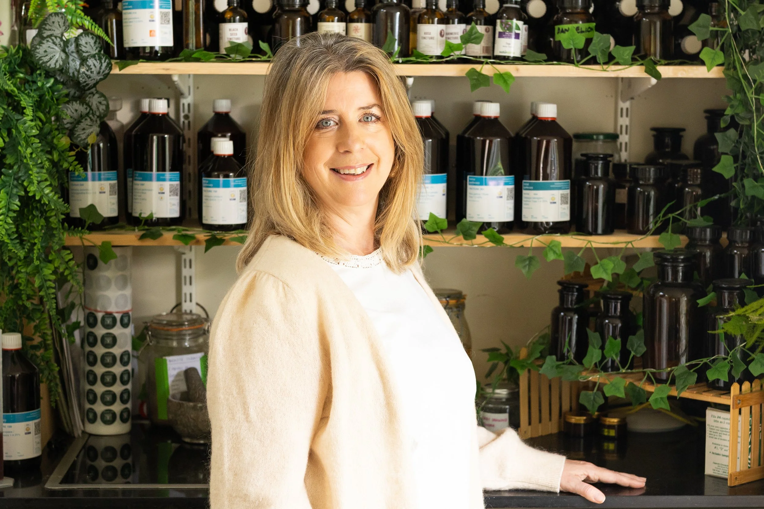 A woman stands in a kitchen or pharmacy surrounded by shelves filled with brown bottles and jars, some with white labels. Green leafy plants decorate the space, creating a natural environment.