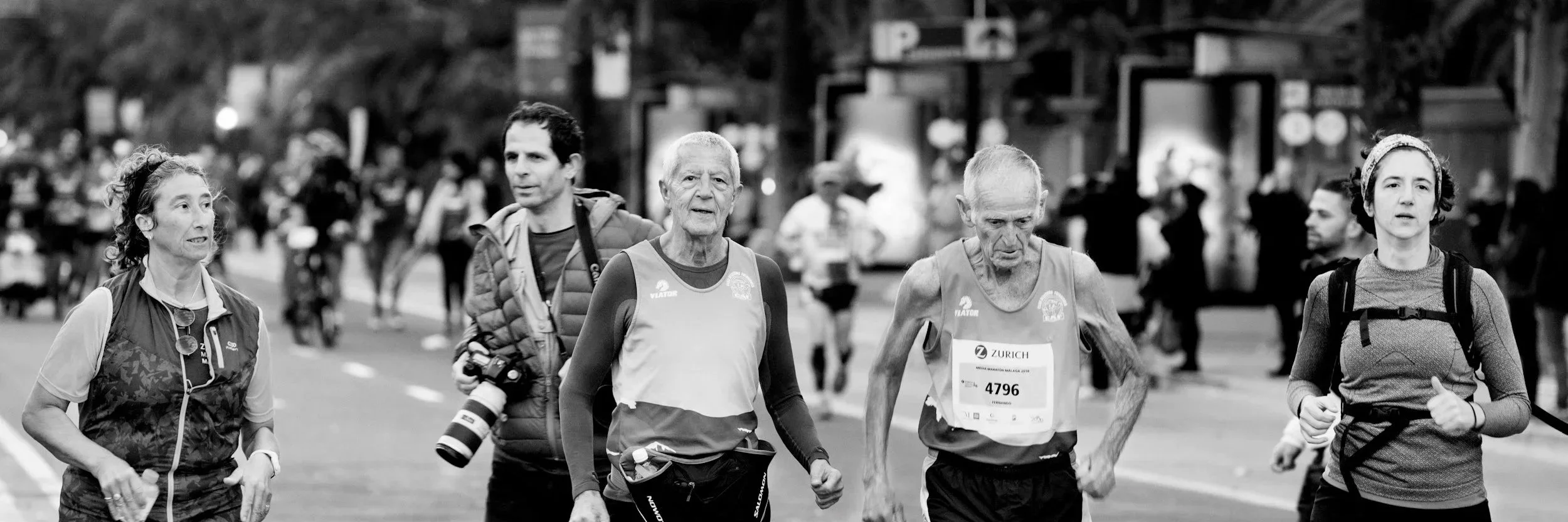 Menschen laufen bei einem Marathonlauf auf einer Stadtstraße, einige tragen Laufkleidung und Nummern, im Hintergrund sind andere Läufer und Stadtbäume sichtbar.