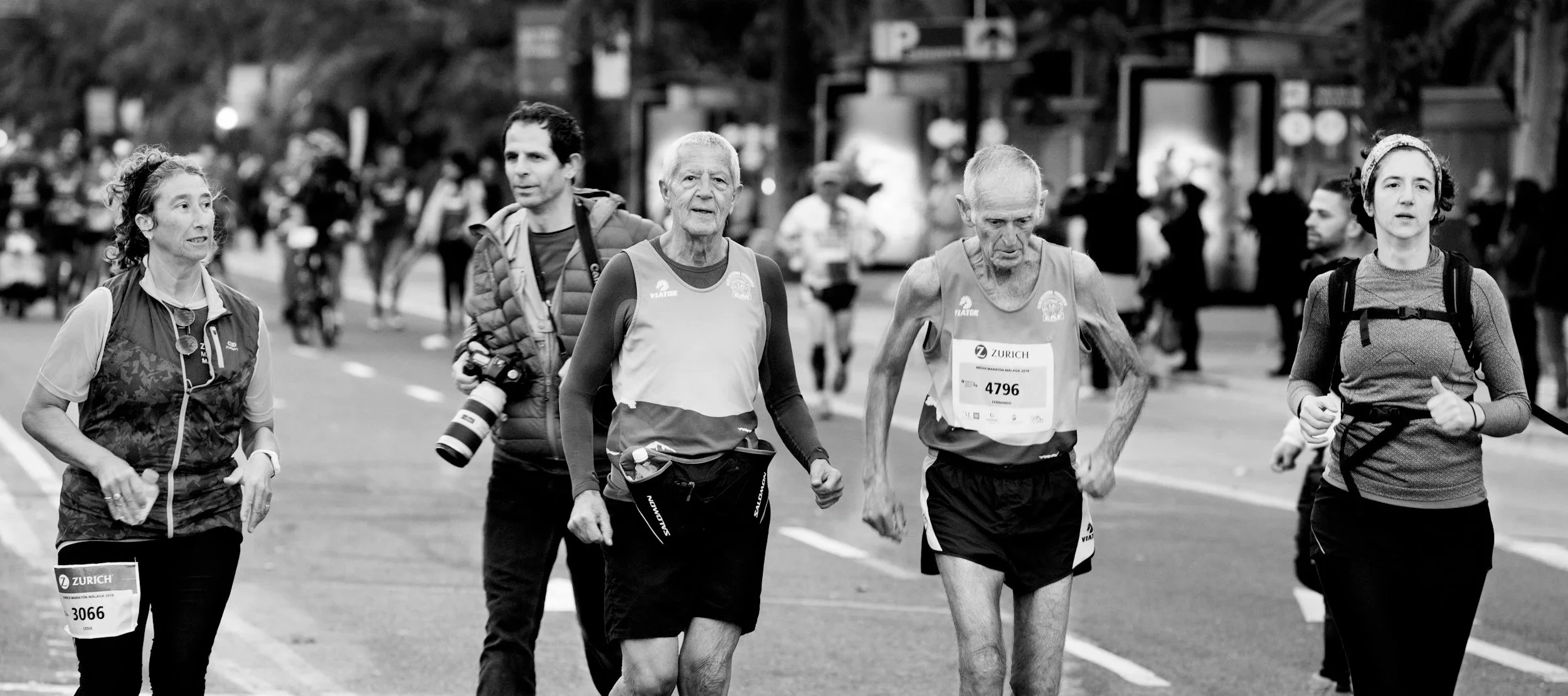 Eine Gruppe von Läufern bei einem Marathon, unterwegs auf einer Stadtstraße, einige mit Startnummern, im Hintergrund sind andere Läufer und Zuschauer.