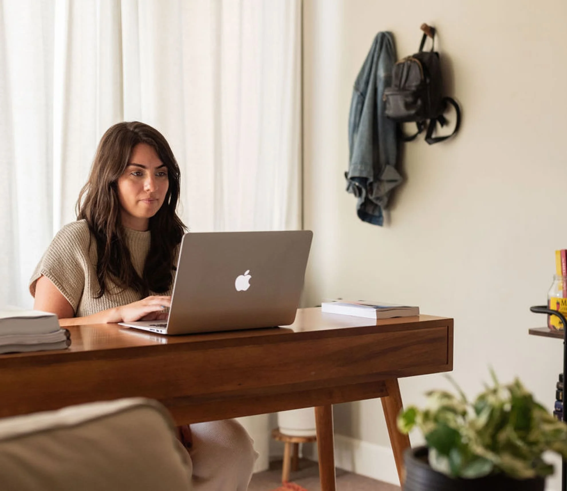 Woman using a silver MacBook laptop at a wooden desk in a well-lit room with beige walls and curtains, with notebooks and a plant nearby.