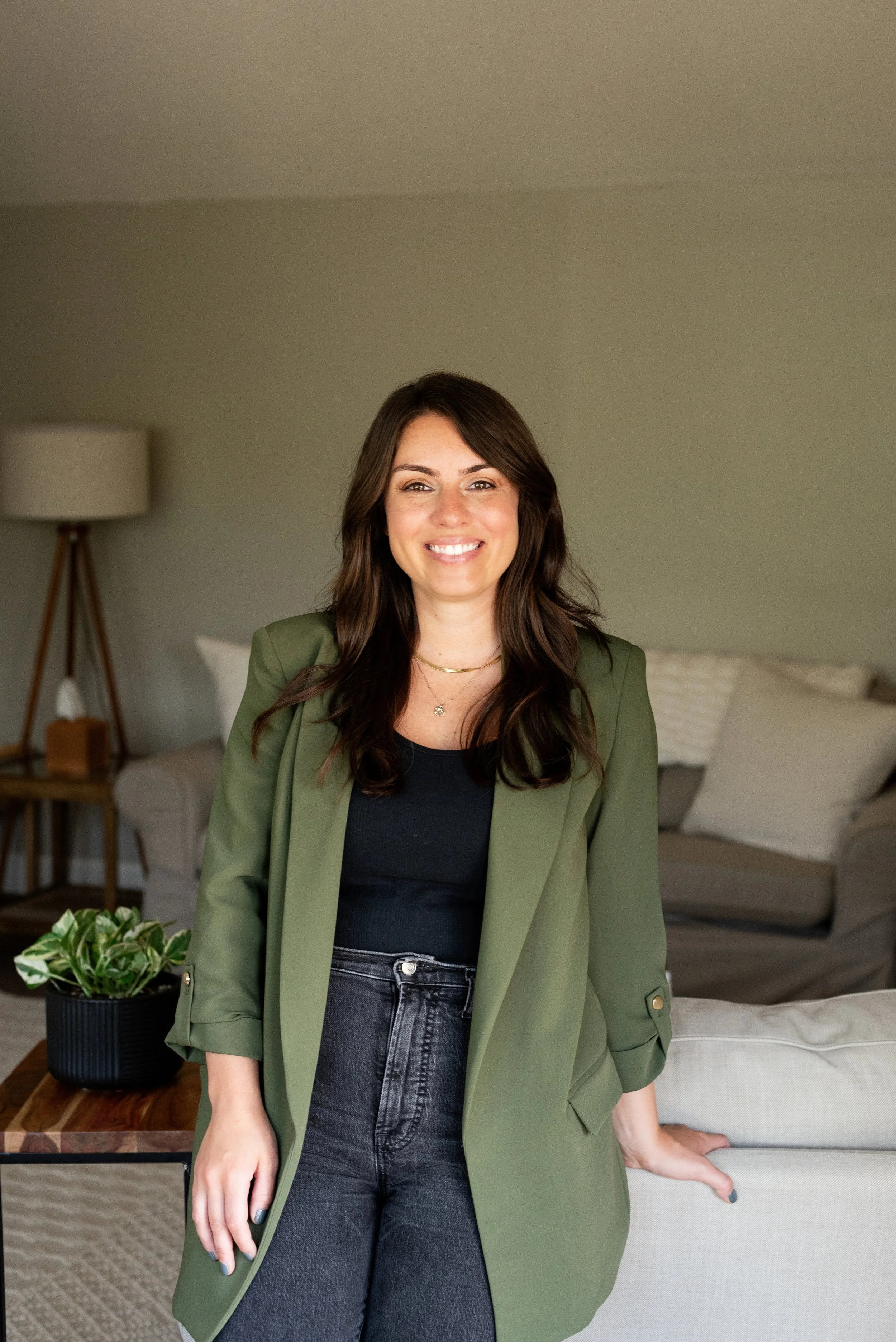 A woman with long dark hair smiling and wearing a black shirt and an olive green blazer, standing in a contemporary living room with a beige sofa, a wooden side table with a potted plant, and a standing lamp.