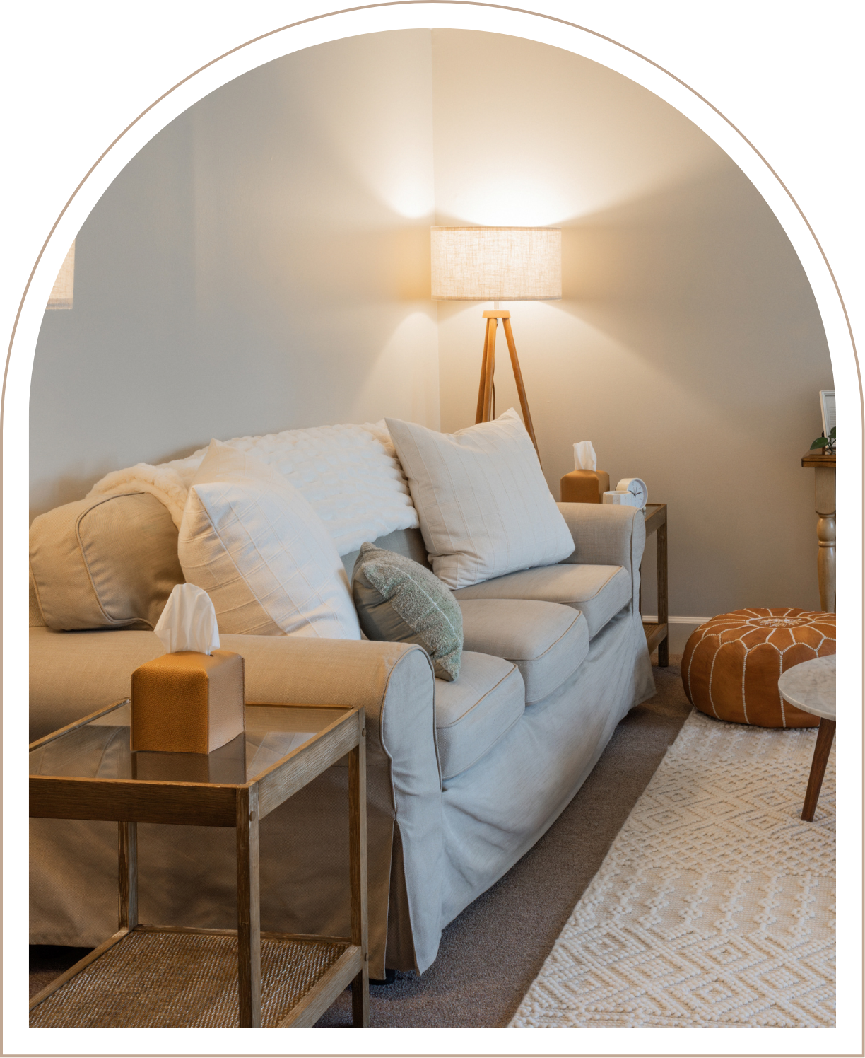 A cozy living room with a beige sofa adorned with multiple pillows, a wooden side table with a tissue box and clock, a tall floor lamp with a beige shade, a small side table, and a round tufted ottoman, beige and white patterned rug on the floor.
