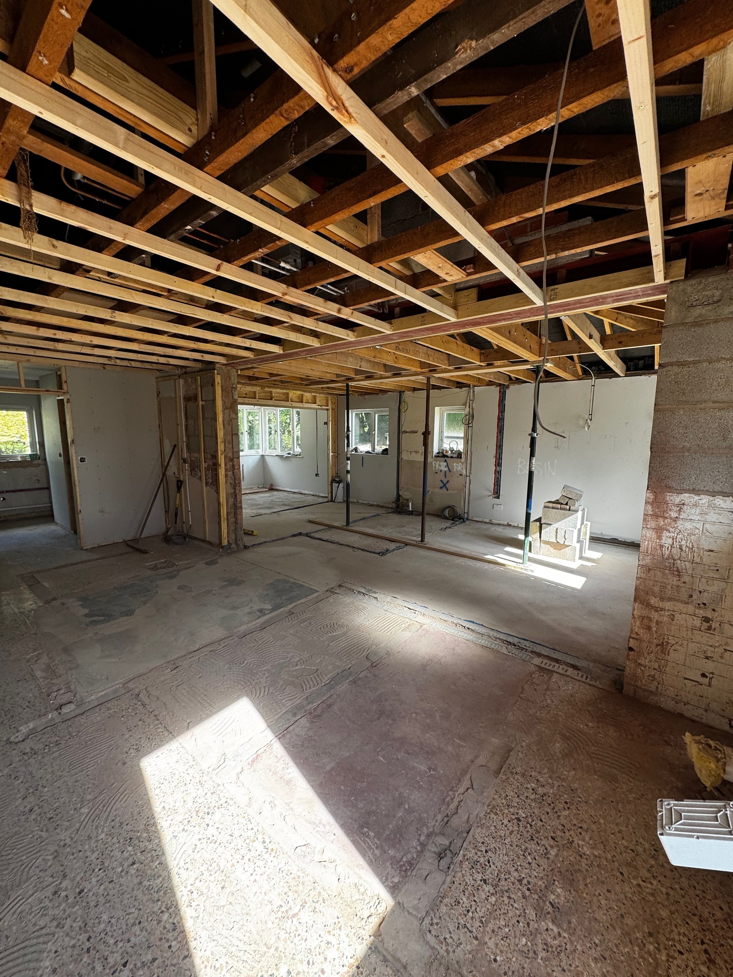 Interior of a house under construction, with exposed ceiling joists, plywood framing, and a partially finished concrete floor.