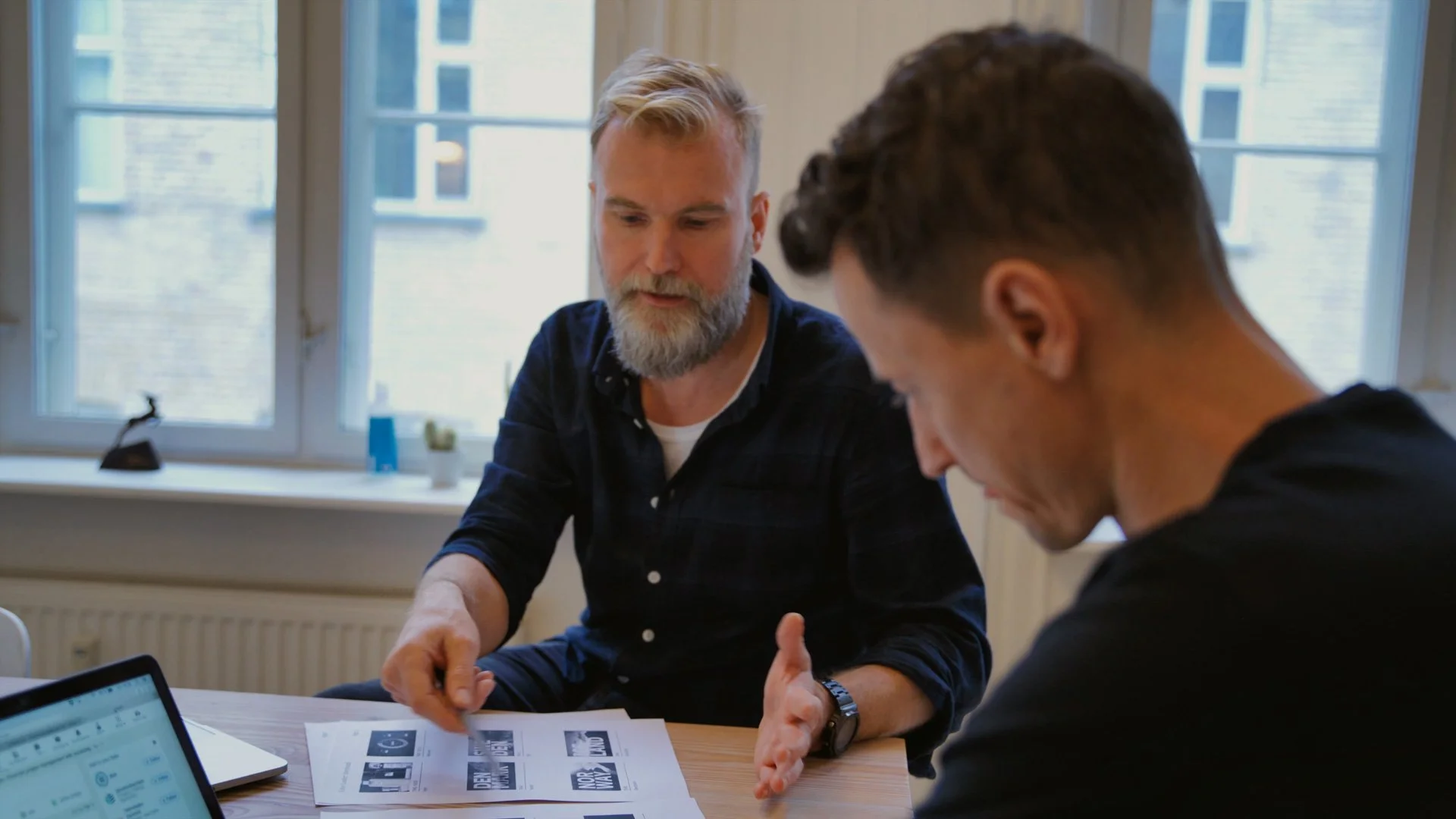 Two men having a discussion at a table, with papers and laptops in front of them, in a room with large windows.