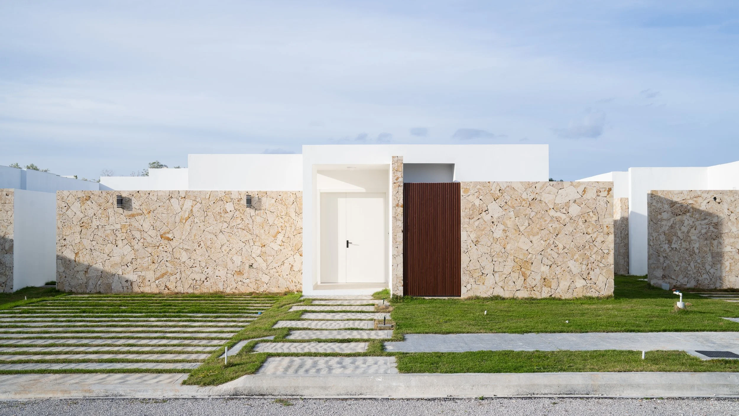 Modern house with white walls, stone accents, and a wooden door, surrounded by a manicured lawn and stone pathway.