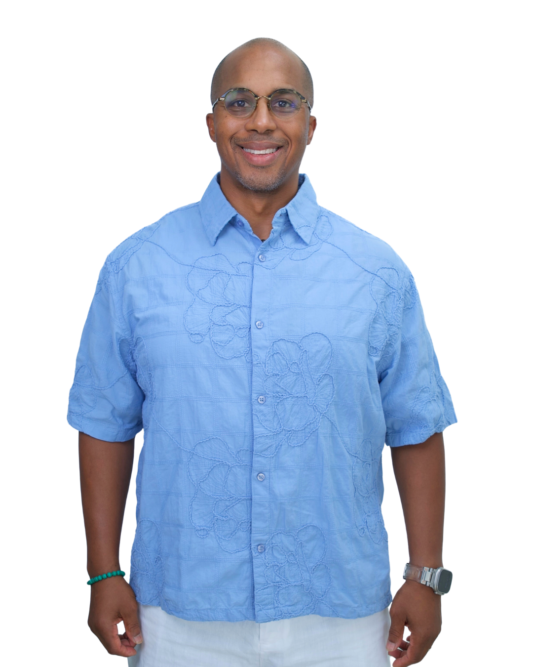 A smiling man wearing a light blue embroidered short-sleeve shirt, glasses, a wristwatch, and a bracelet, standing against a white background.