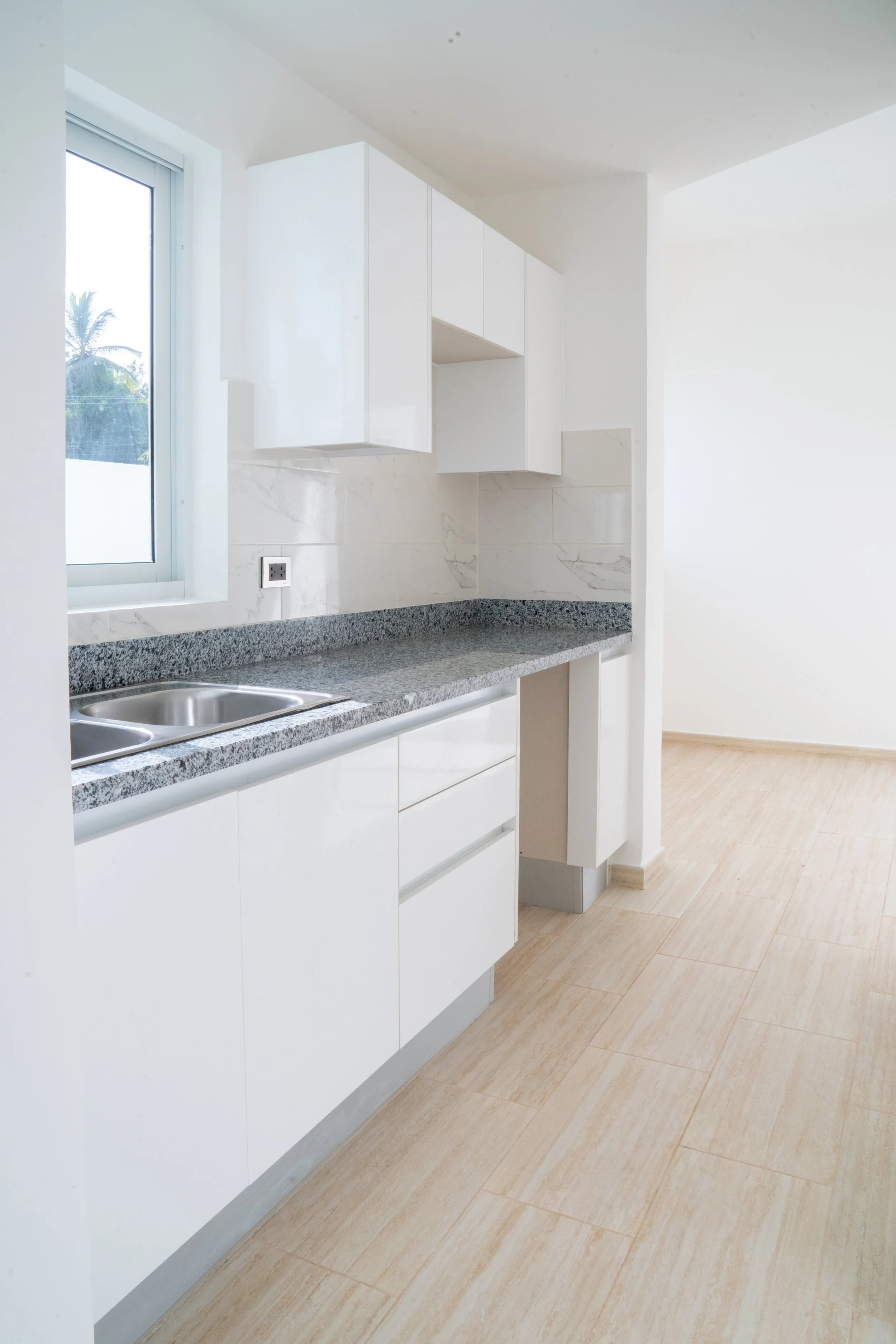 Empty kitchen with white cabinets, granite countertop, sink, window, and beige tile flooring.