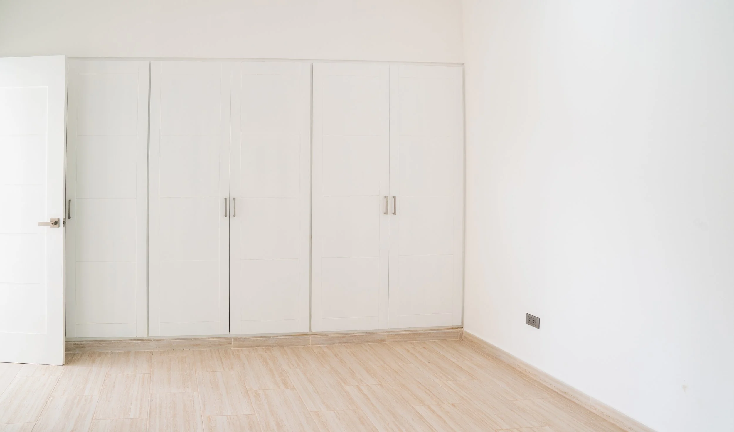 Empty white room with built-in white closet and a light wood floor, electrical outlet on the right wall.
