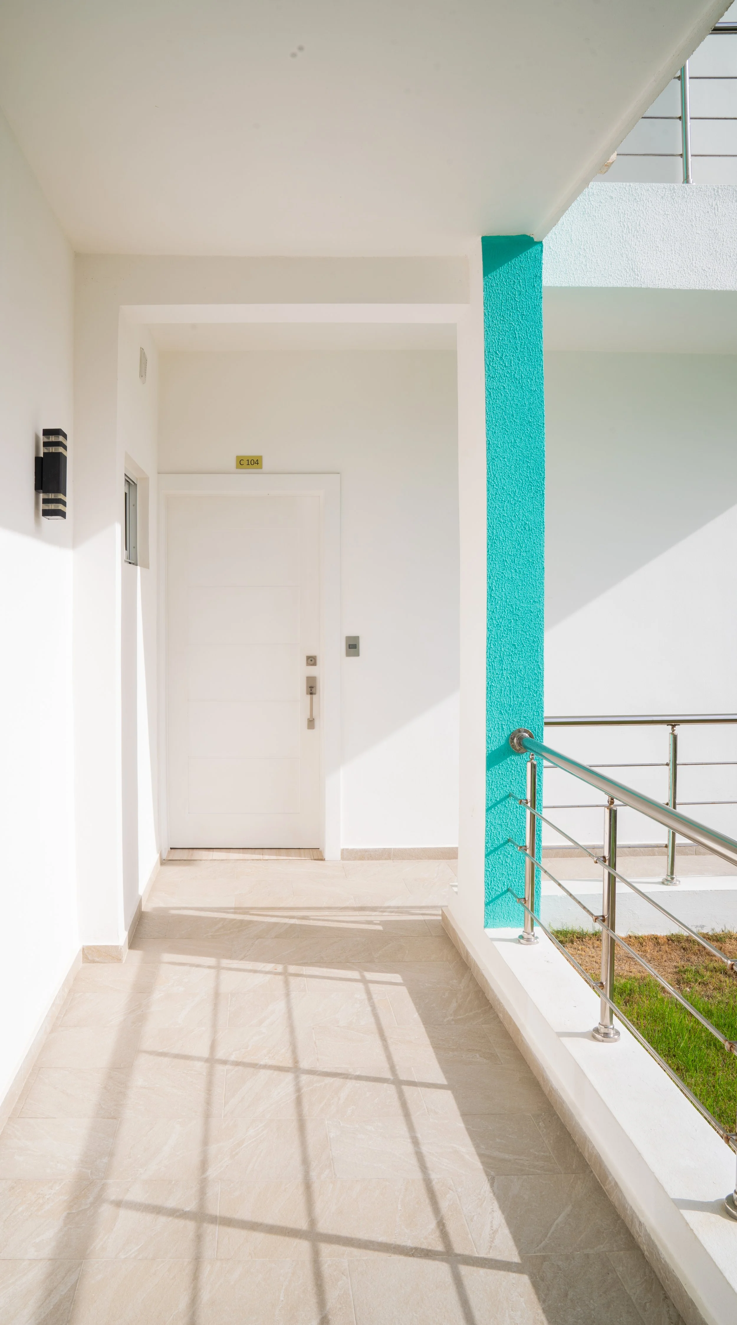 Exterior hallway with white walls, beige tiled floor, teal accent wall, stainless steel railing, and white front door with gold room number sign C 104.