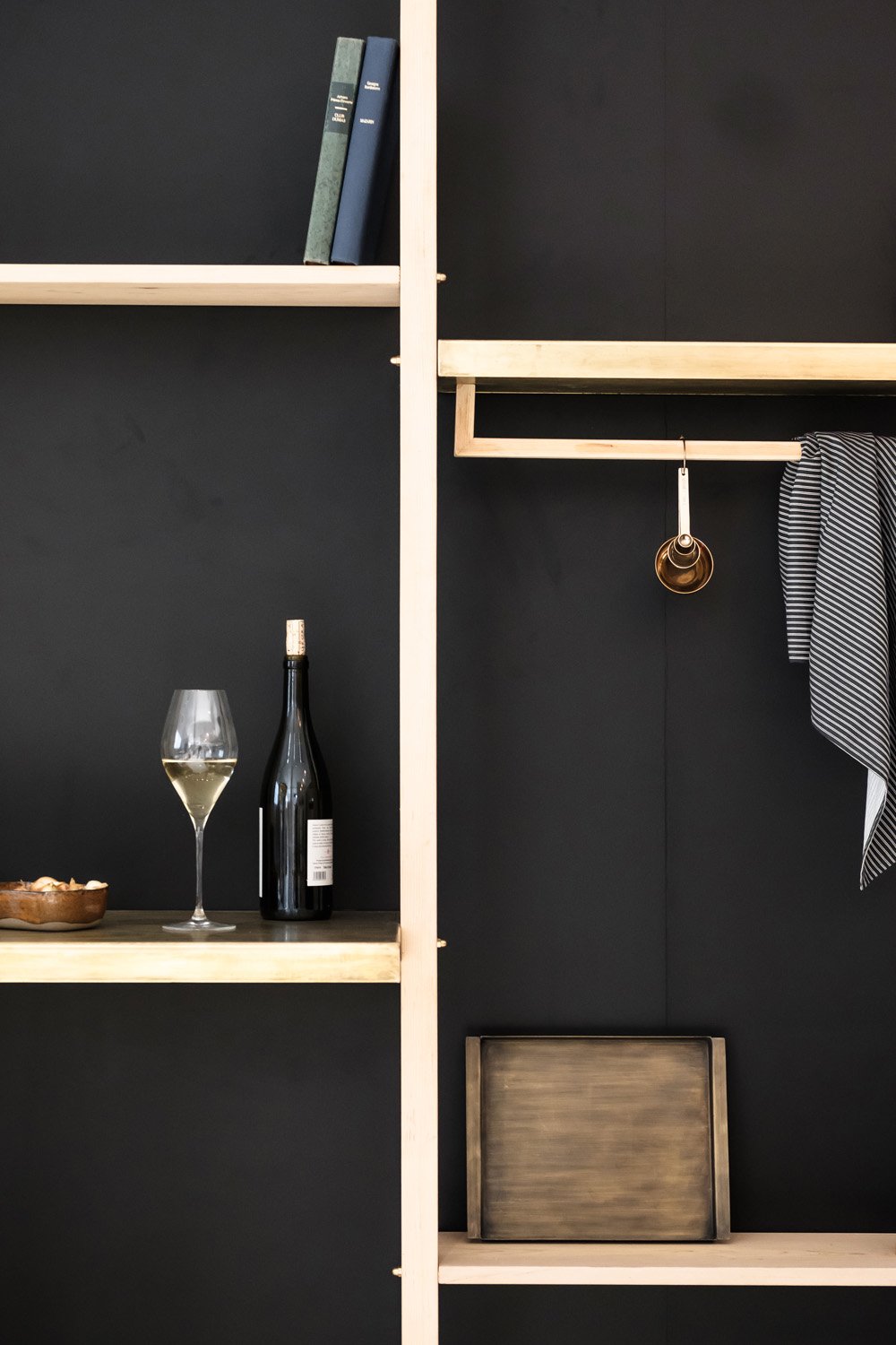 A modern shelving unit with black backing and light wooden frame, holding books, a wine glass with white wine, a wine bottle, a wooden tray, a hanging set of small golden cups, a striped cloth, and a rectangular wooden box.