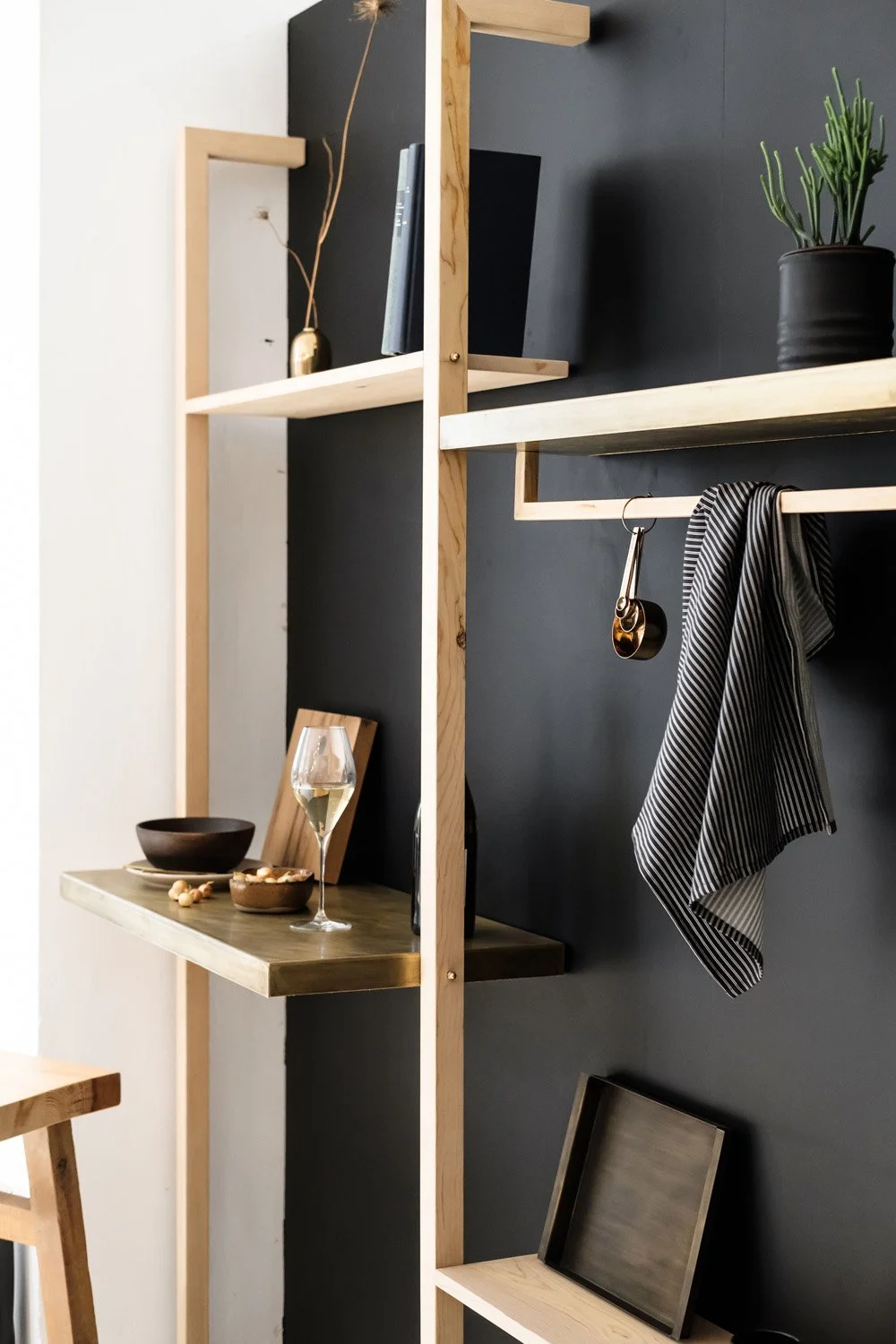 Interior view of a modern room with wooden shelves and a black chalkboard wall, holding a plant, a wine glass, a bowl, and picture frames, with a striped cloth hanging on a hook.