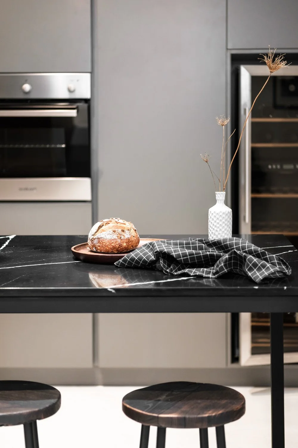 A kitchen with a black marble table, a loaf of bread on a round plate, a black and white checkered cloth, and a white vase with tall dried plants.