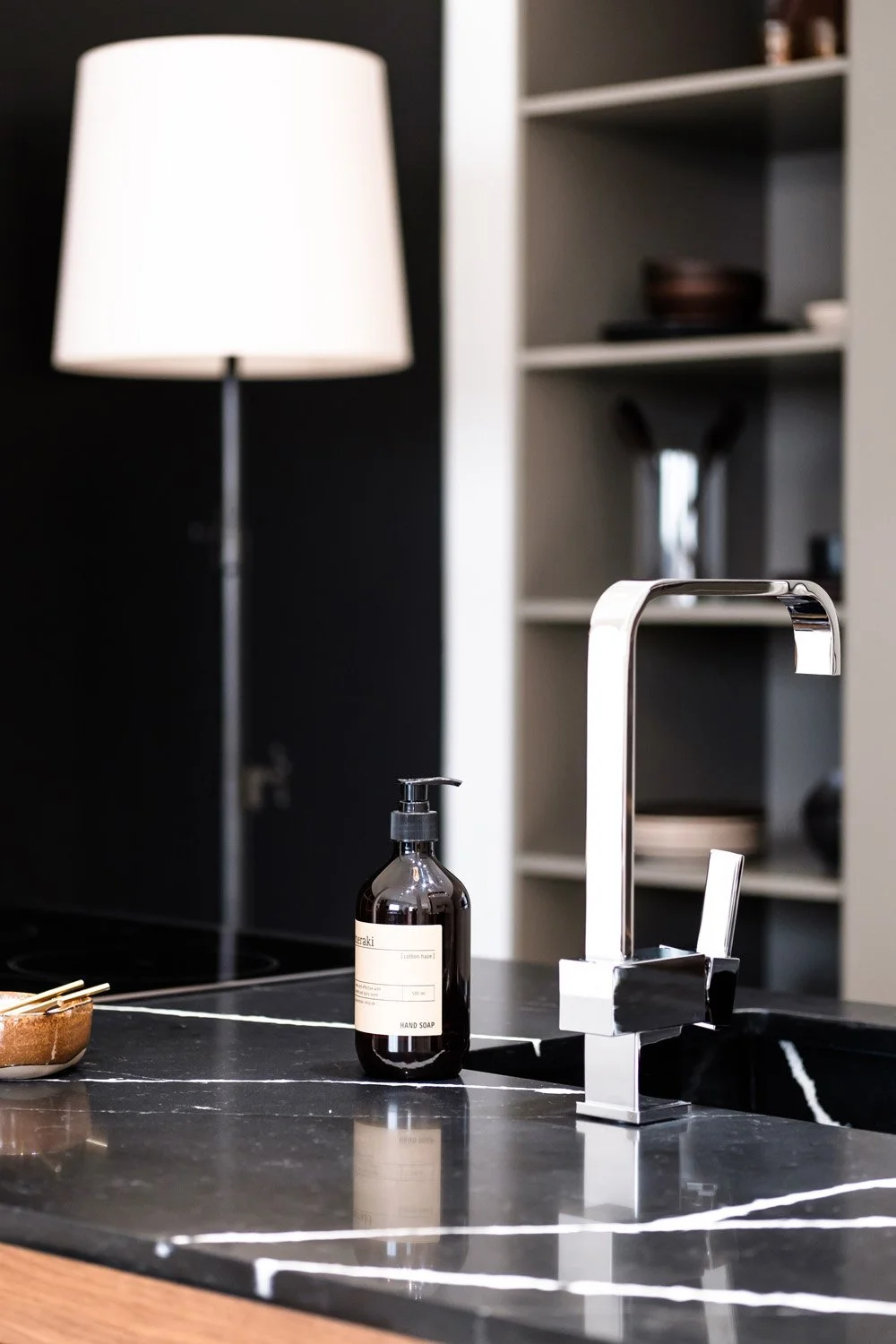 Modern kitchen sink with chrome faucet, black soap dispenser, and a small brown bowl on a black marble countertop, with a white lamp and a white shelf in the background.