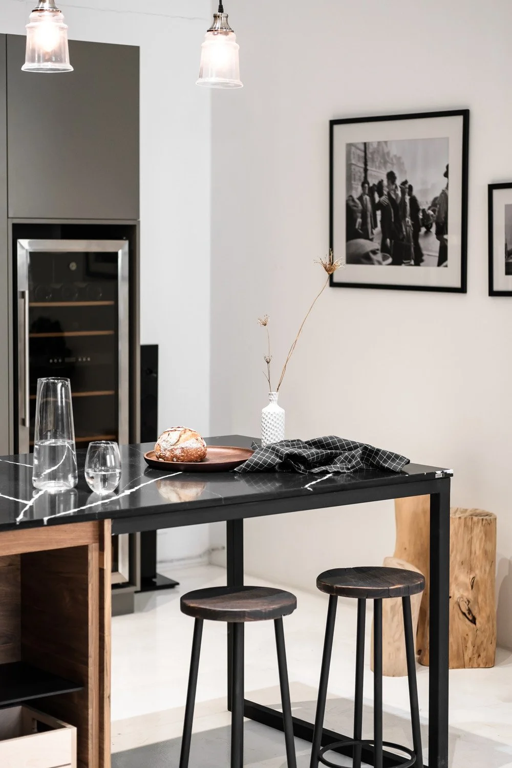 Modern dining area with black and white decor. A black marble table has a glass carafe, two glasses, a bread loaf on a copper plate, and a black checked napkin. Two black stools surround the table. On the wall are two black and white photographs. The