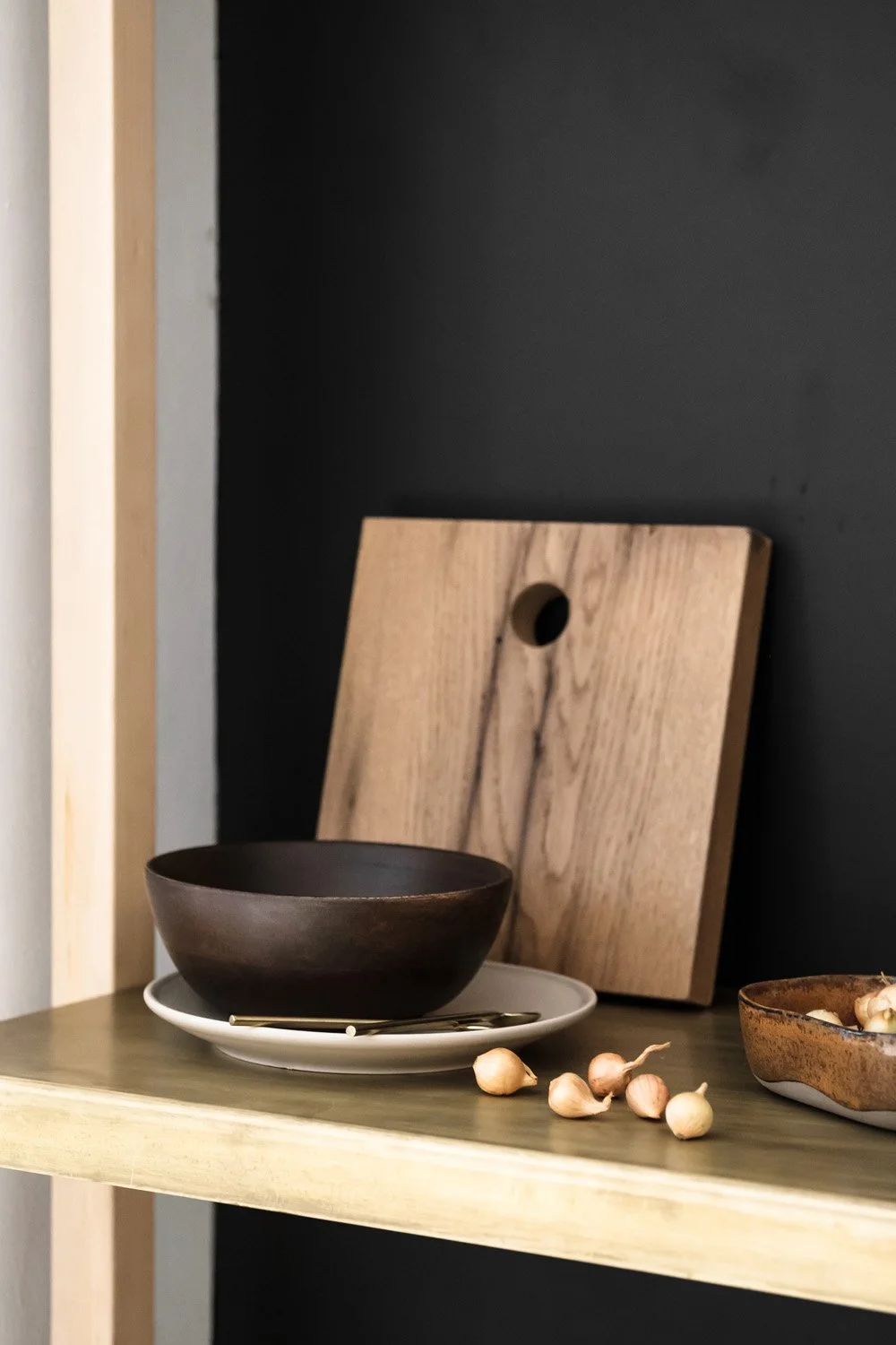Dark wooden bowl on a white plate with a gold utensil underneath on a wooden surface, beside a small bowl of garlic cloves and a wooden cutting board in front of a dark wall.