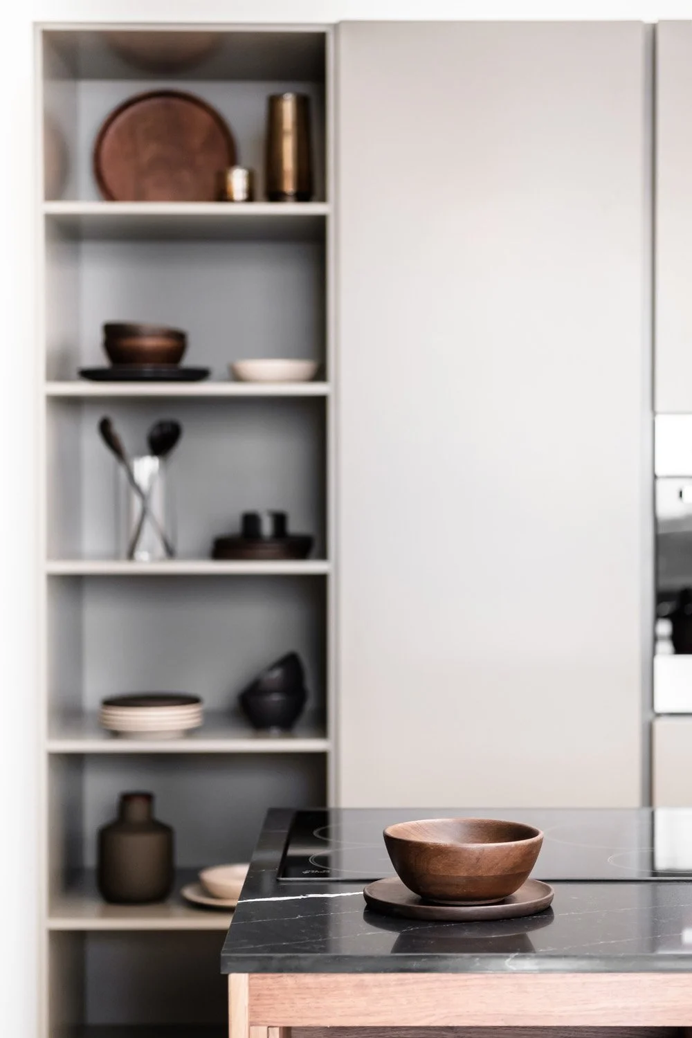Wooden bowl on a plate on a black countertop in a modern kitchen, with open shelves holding dishes and decorative items in the background.