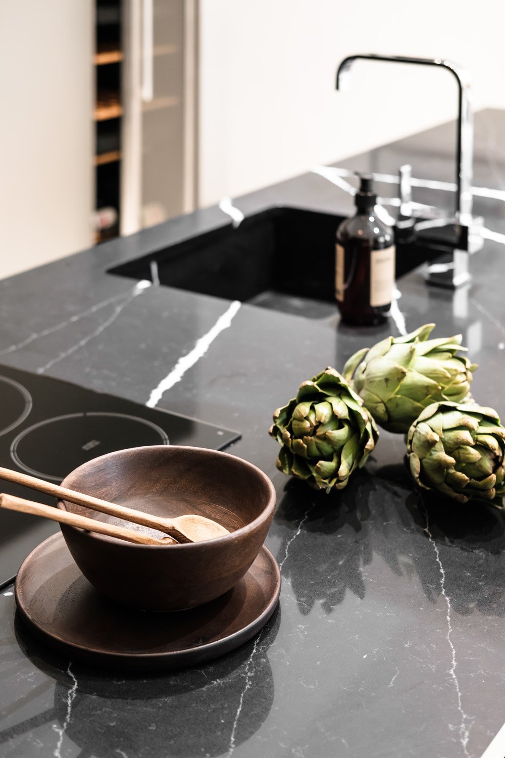 Black marble kitchen countertop with Artichokes, wooden bowl and utensils, soap dispenser, and electric stove. Modern kitchen design.