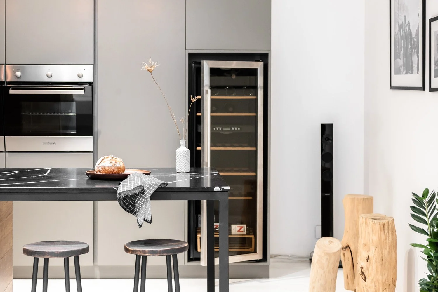 Modern kitchen with black marble island, two stools, a bread plate, vases, a wine fridge, and wooden stools.