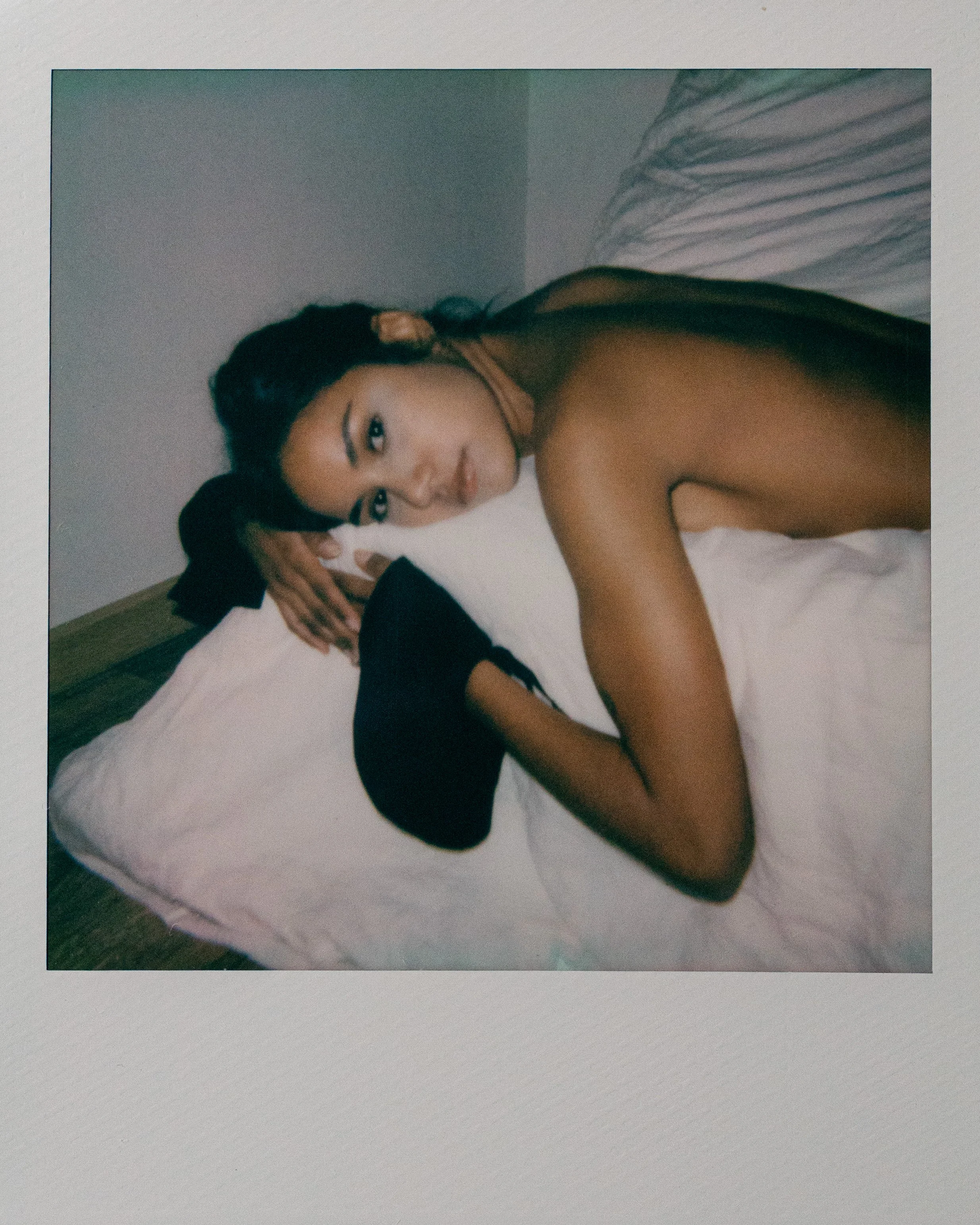 A woman lies on her side on a bed, resting her head on a white pillow, looking at the camera, with a black hat in front of her.