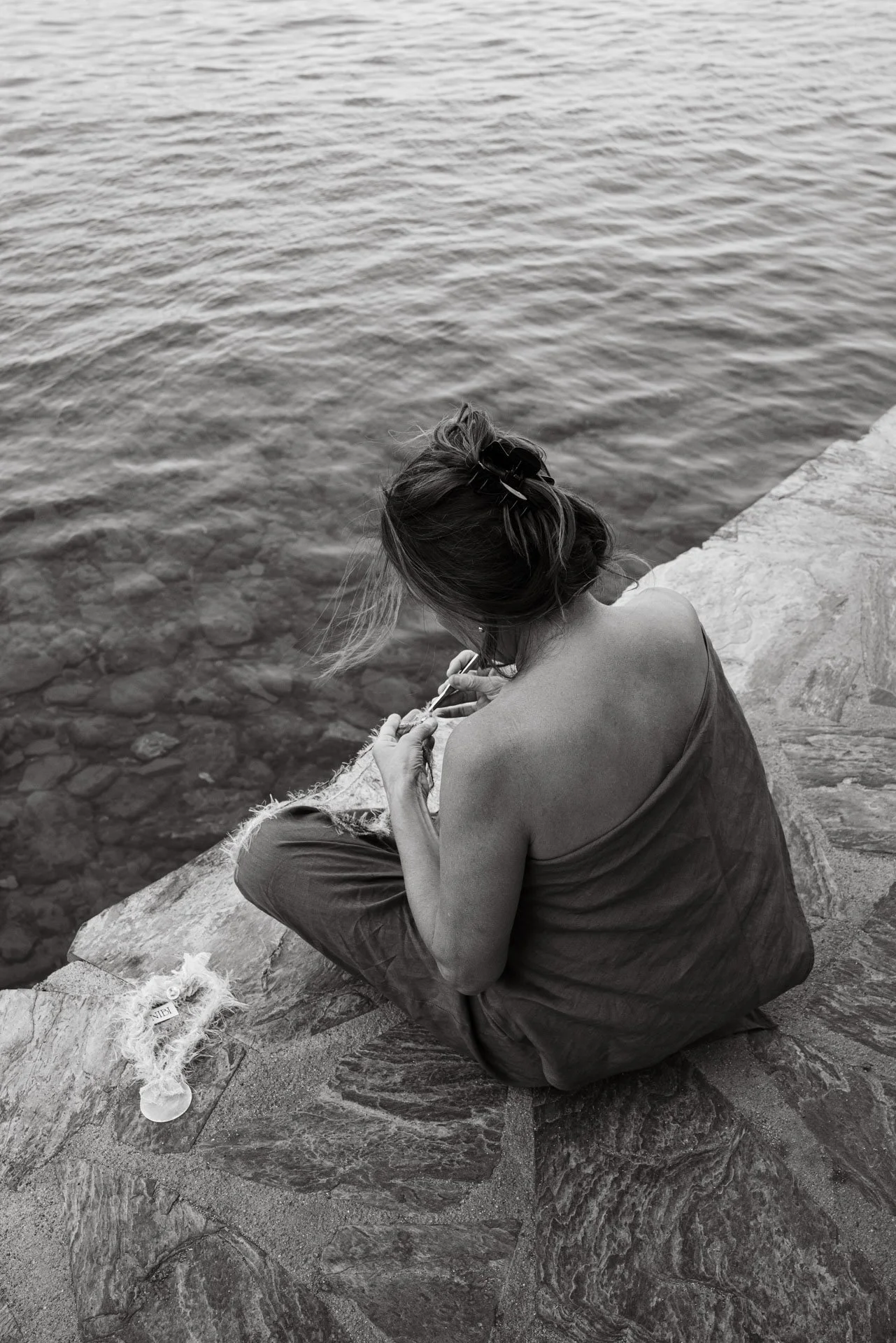 A woman sitting on a stone edge by the water, sewing or knitting with a needle and thread, with a bottle and some fabric nearby.