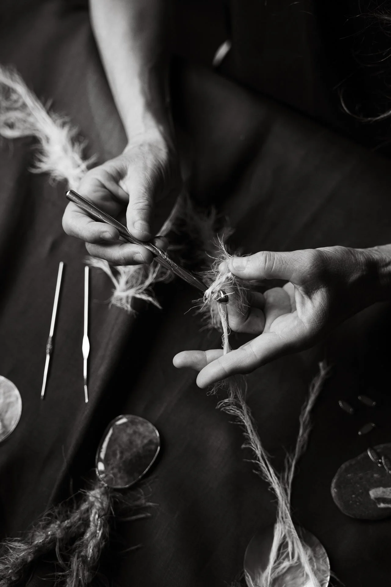 A person working on a handmade necklace, holding a strand of yarn in one hand and manipulating a knitting needle with the other, with various crafting tools and mirrors on the table.