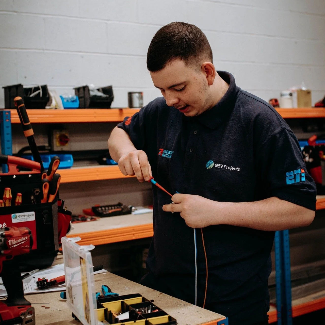 A man working on an electronic project in a workshop, with tools and equipment on shelves and a workbench.