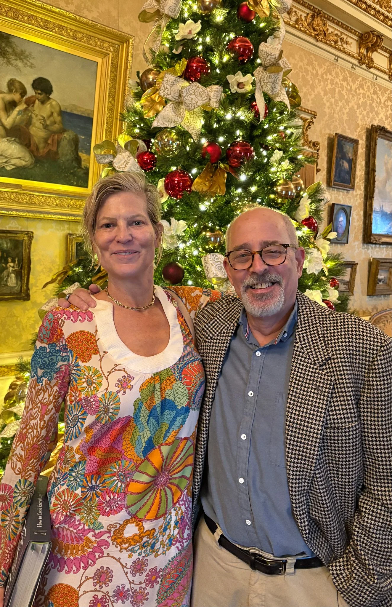 A smiling woman and man standing together in front of a decorated Christmas tree, with framed paintings on a wall behind them.