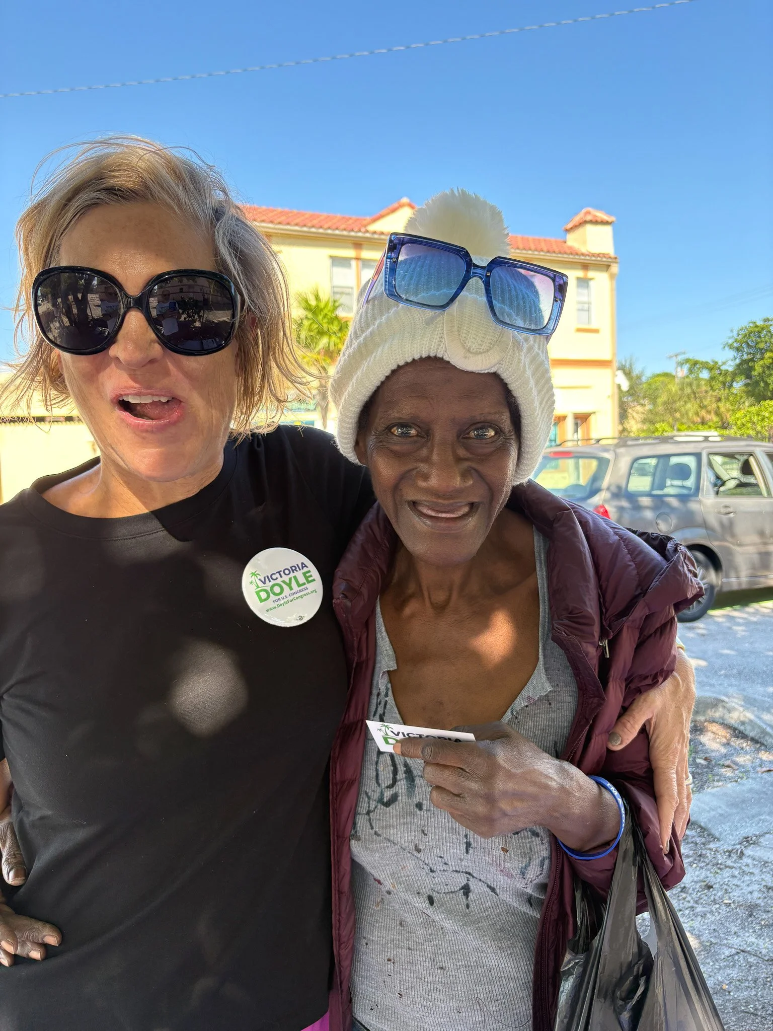 Two women smiling outdoors on a sunny day, one wearing dark sunglasses, a black shirt with a campaign button, and the other wearing a white beanie with sunglasses on top, and a maroon jacket, holding a flyer, with houses and parked cars in the backgr
