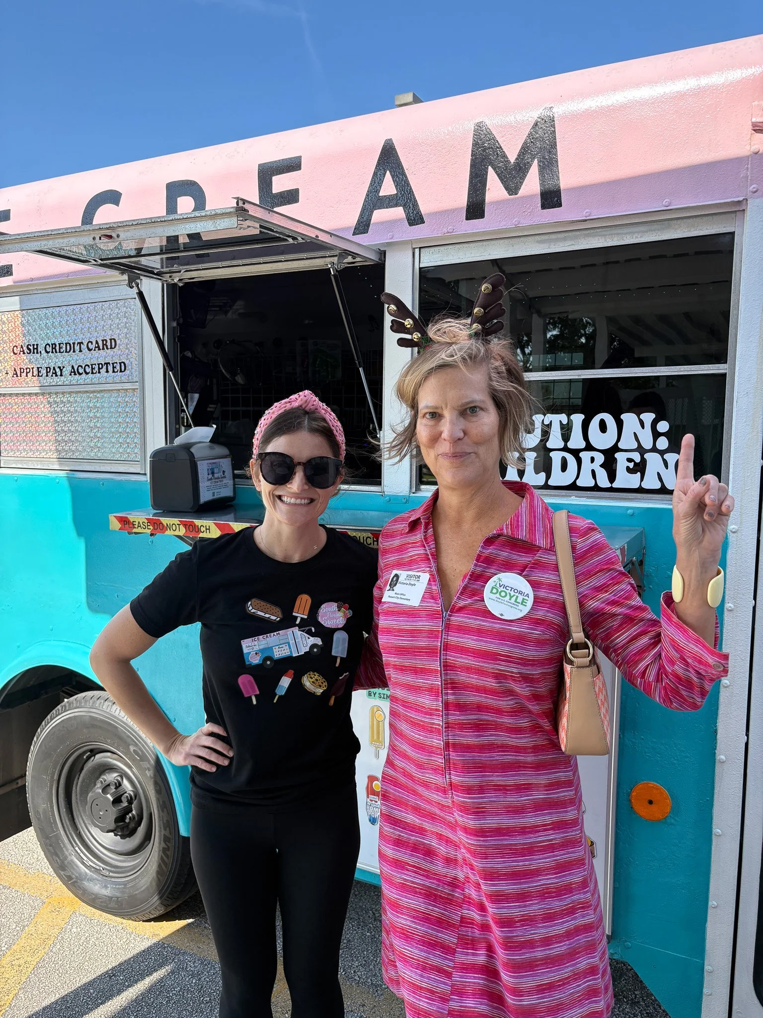 Victoria Doyle and constituent in front of a colorful ice cream truck, one wearing reindeer antlers and the other in sunglasses and a black shirt with ice cream illustrations.