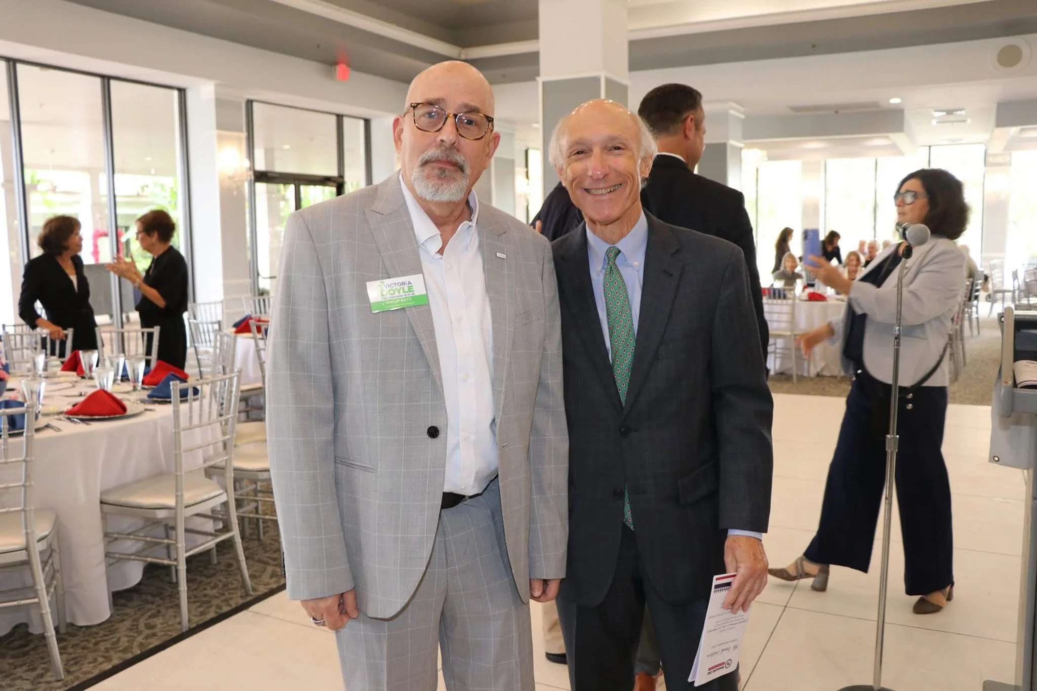 Philip Katz and a man in suits standing together at an indoor event, smiling for the camera. One has a name tag that reads 'Victoria Doyle'. In the background, other people are socializing, with tables set up and a woman taking a photo.