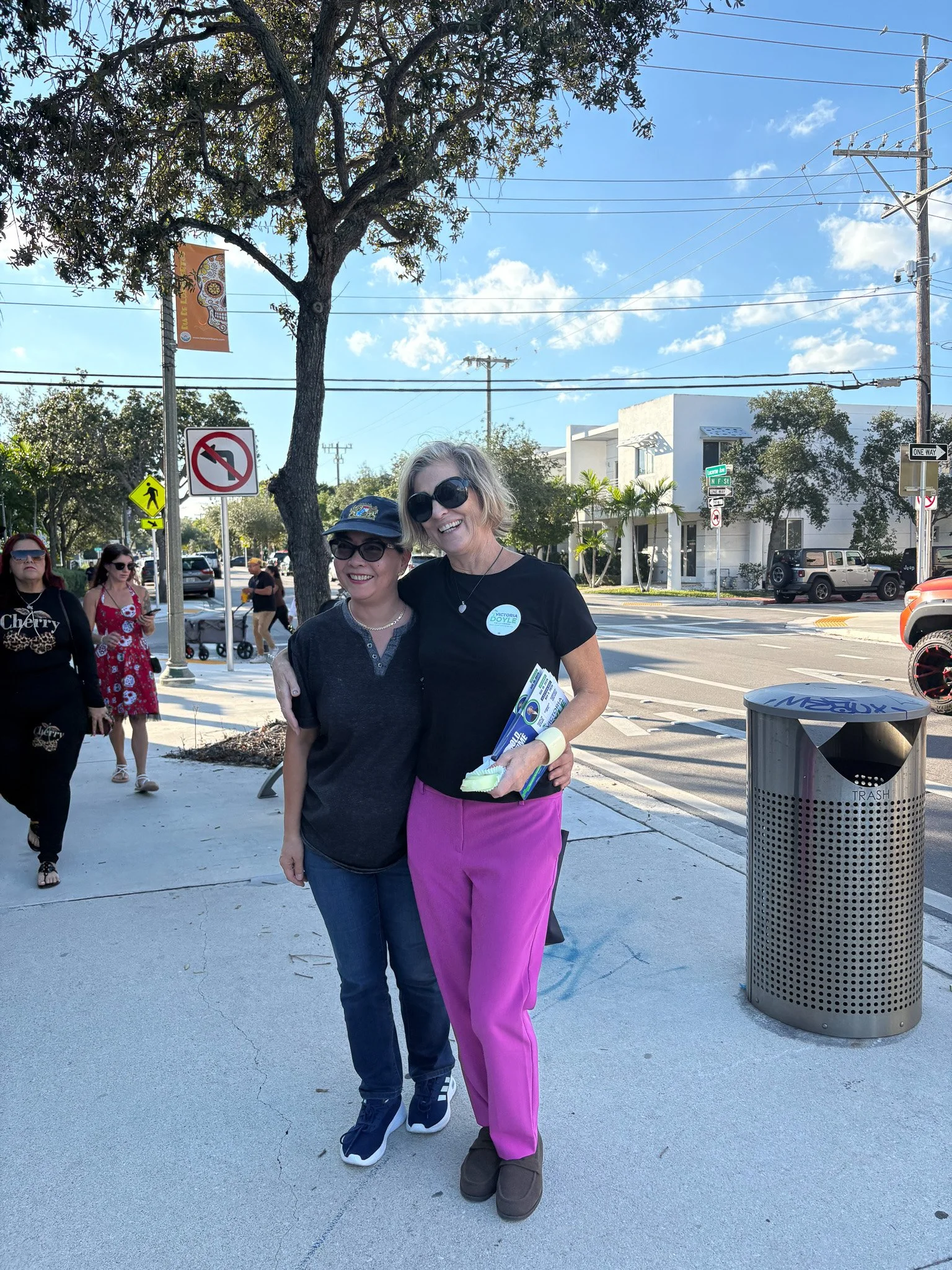 Two women smiling and hugging on a sidewalk, with other pedestrians in the background, a tree, traffic signs, and a trash can nearby.