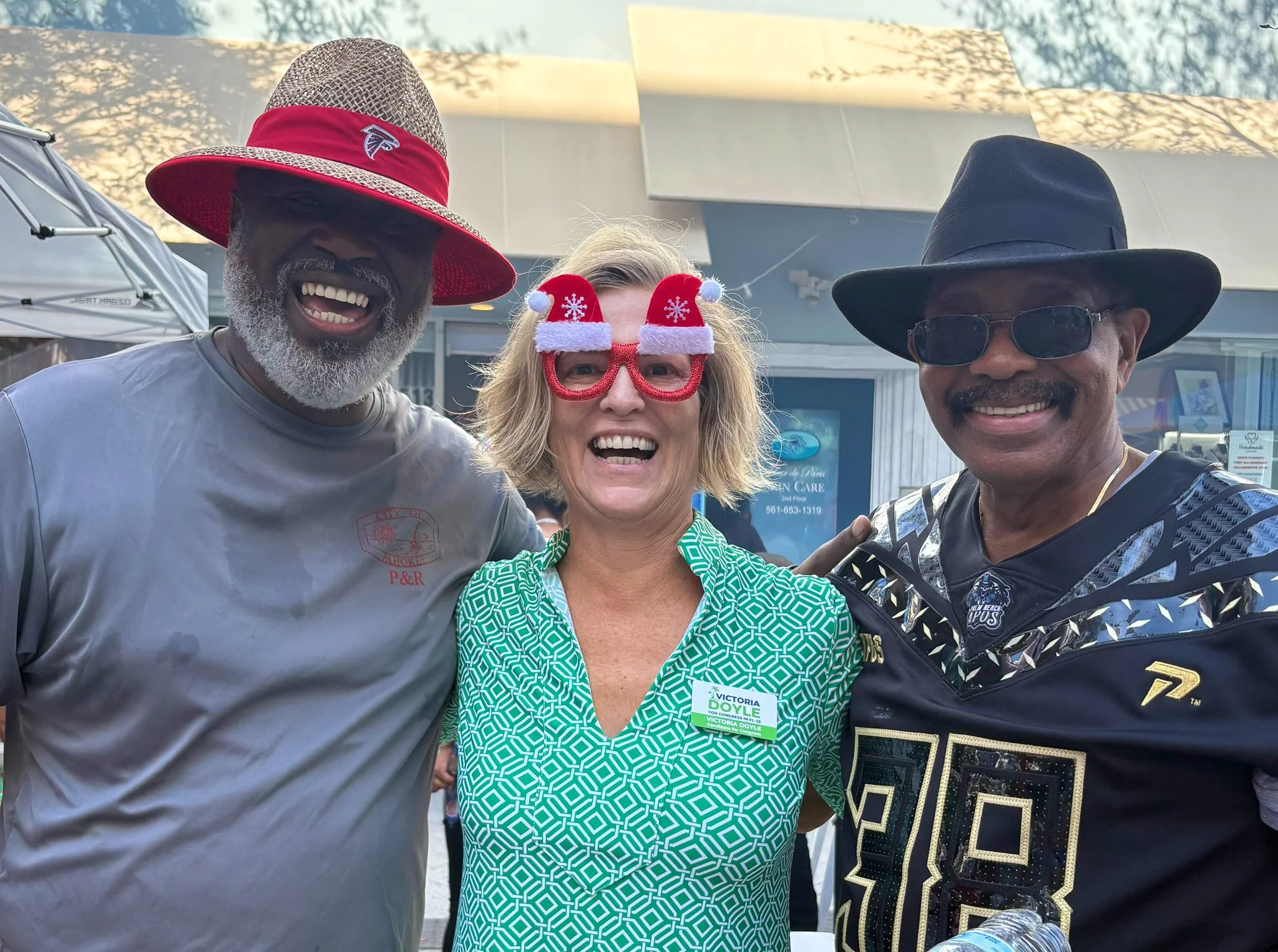 Three people smiling and wearing festive and casual accessories, standing outdoors in front of a building with umbrellas and signs related to healthcare and a phone number.