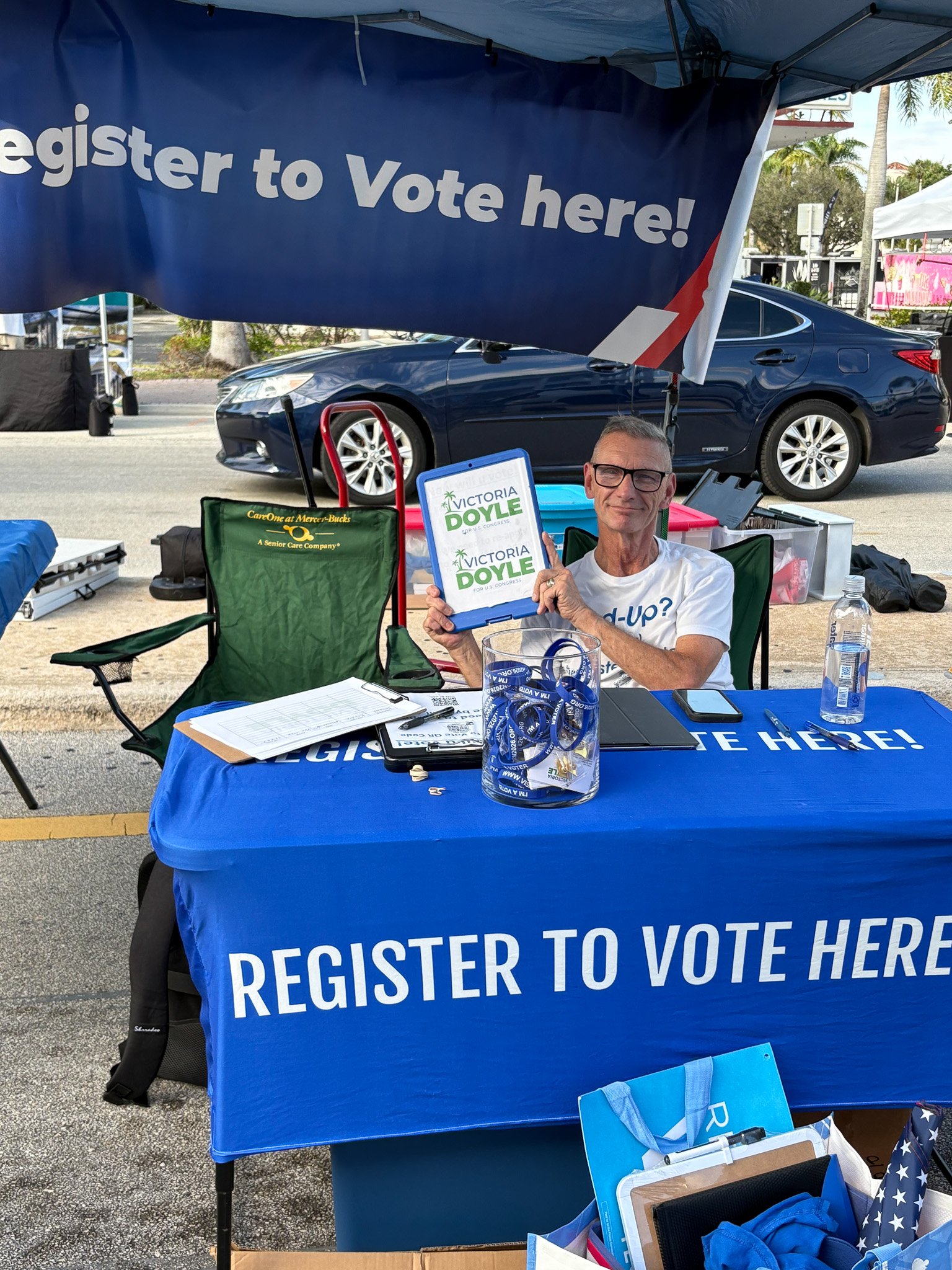 A man sitting at a table under a canopy with a blue banner that says "Register to Vote Here!" He is holding a sign that says "Victoria Doyle for U.S. Congress" and is smiling. The table has registration forms, a water bottle, and other materials.
