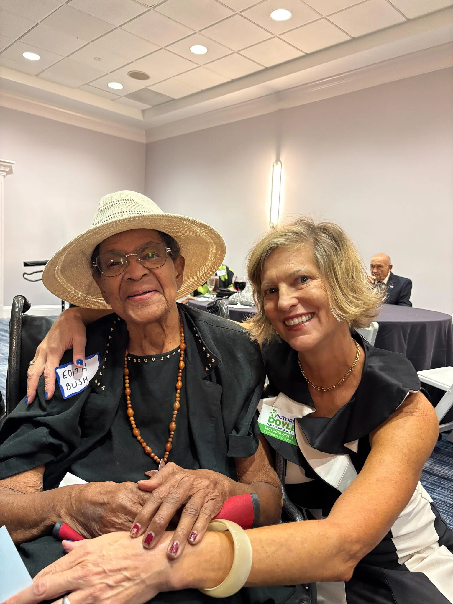 Victoria Doyle with constituent at a conference or event, with one wearing a large straw hat, glasses, and a beaded necklace, and the other wearing a black and white dress and a name badge.