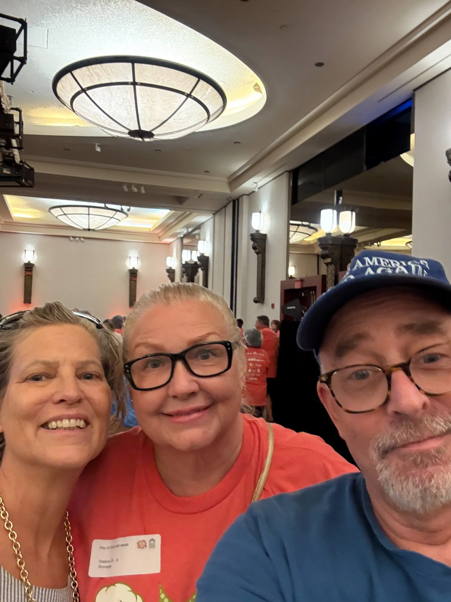 Three people taking a selfie together at an indoor event with a large, well-lit ceiling and numerous attendees in the background.