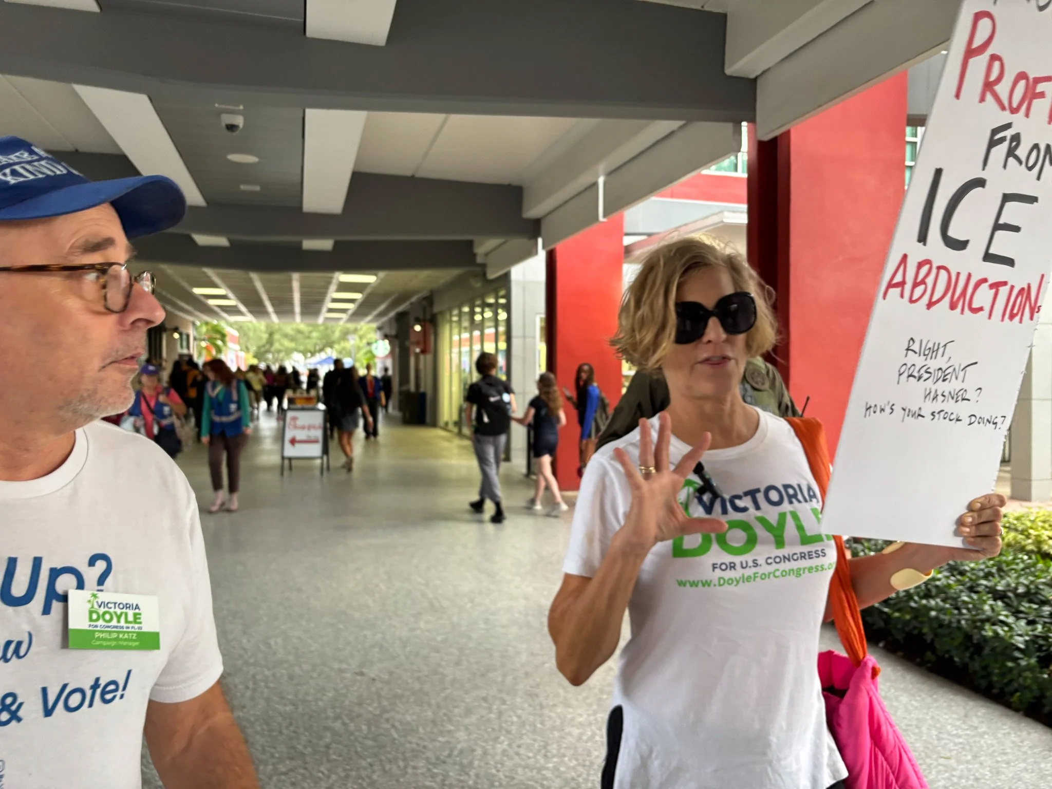 holding a sign that reads 'PROFITEERS FROM ICE ABDUCTION' at a political event, wearing sunglasses and a white T-shirt with the text 'VICTORIA DOYLE FOR U.S. CONGRESS.' She is making a gesture with her hand and has a pink bag over her shoulder. A man