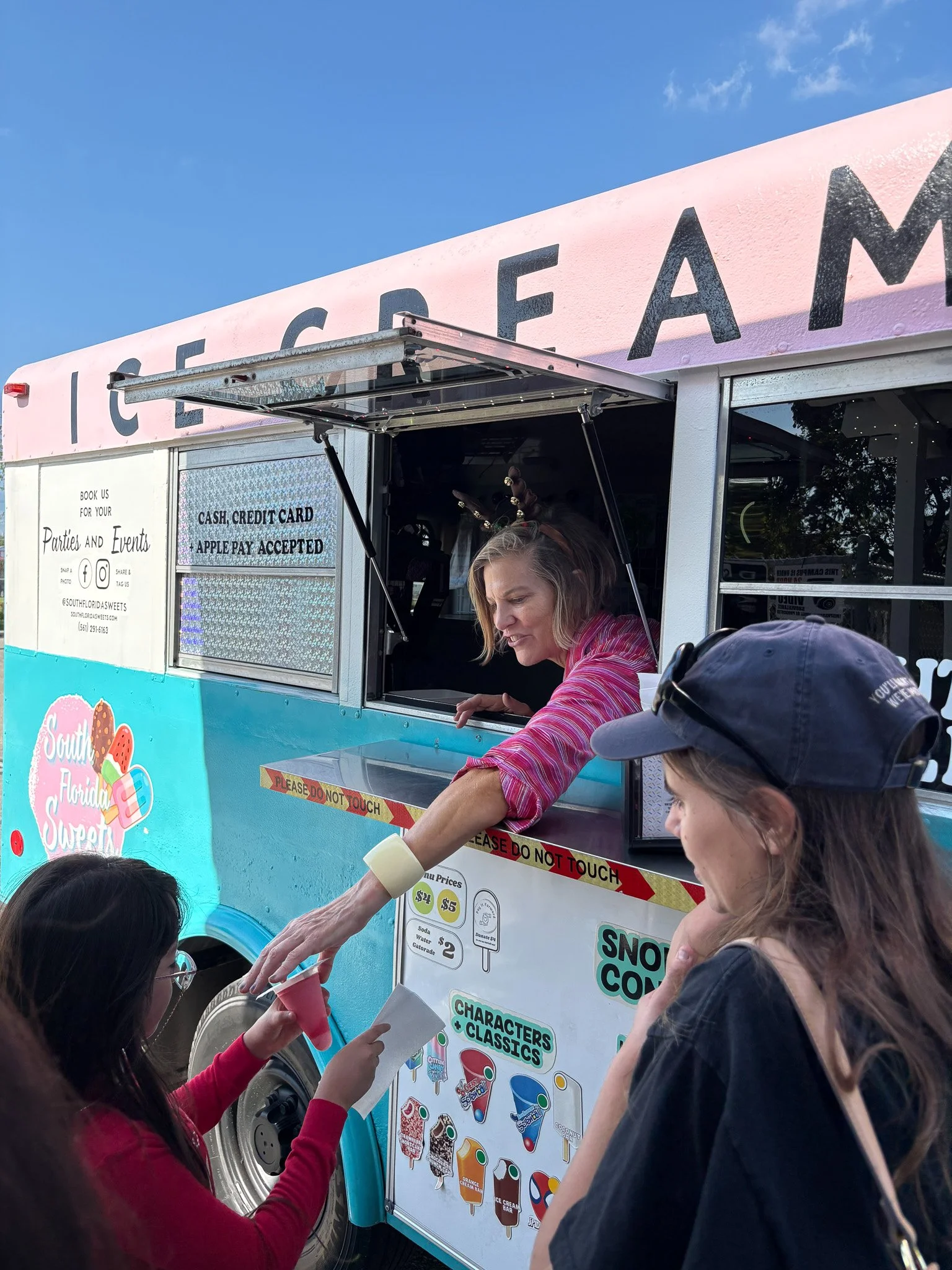 Victoria Doyle in Ice cream truck serving customers with a woman handing ice cream to a customer and a girl in red holding a cone, in front of the truck on a sunny day.