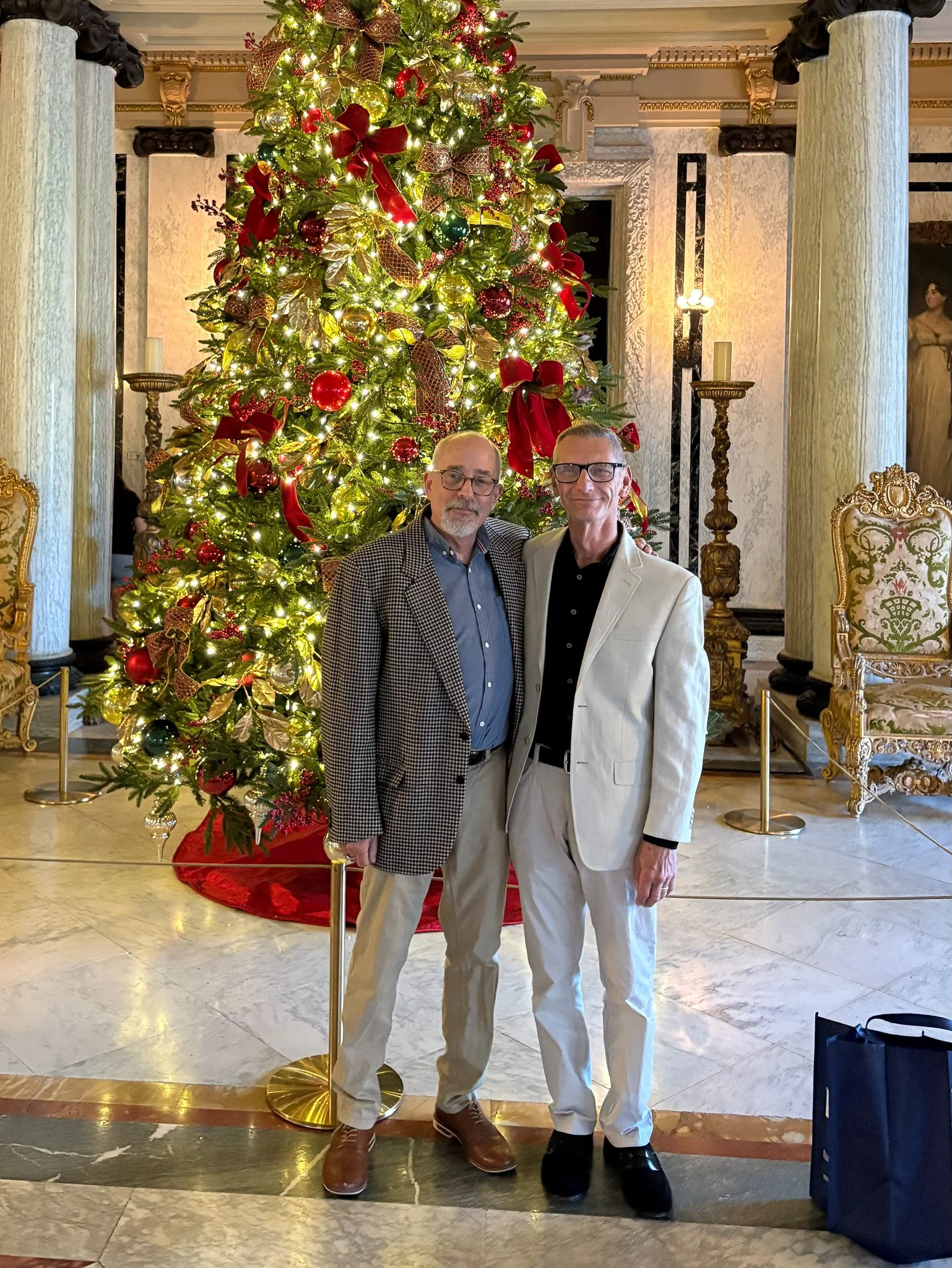 Two men standing in front of a decorated Christmas tree inside a luxurious, ornate room, both wearing suits and smiling at the camera.