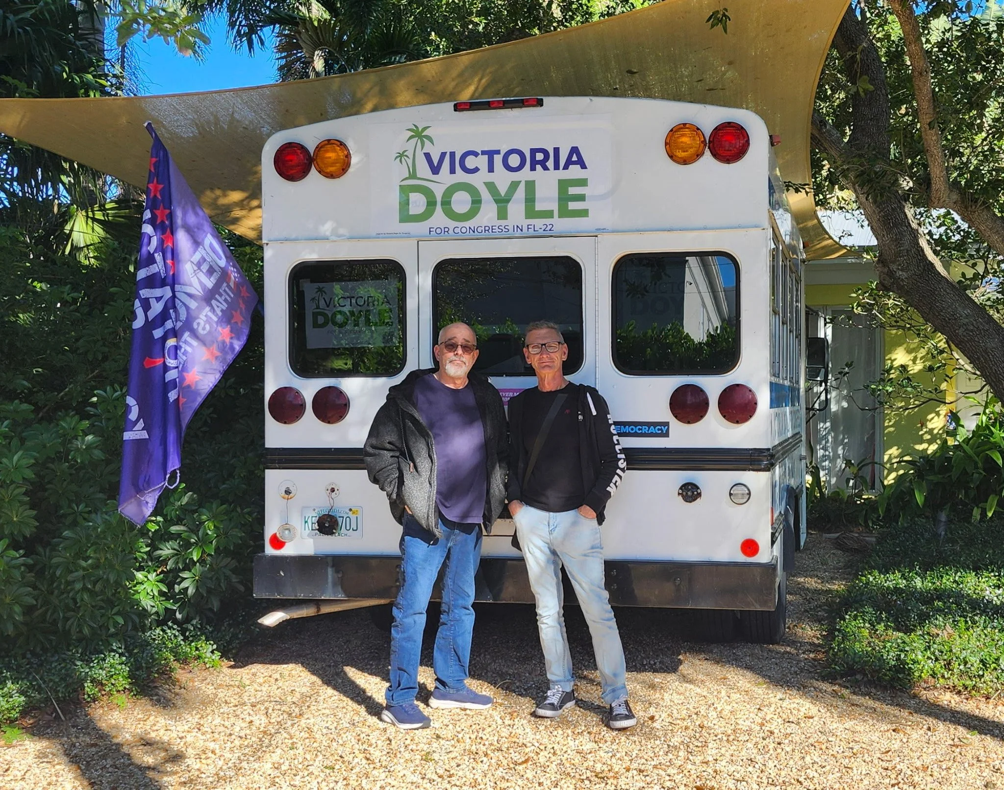 Doyle For Congress bus with 2 men standing in front of a campaign bus with a sign supporting Victoria Doyle for Congress, Florida, 2022, amid greenery.