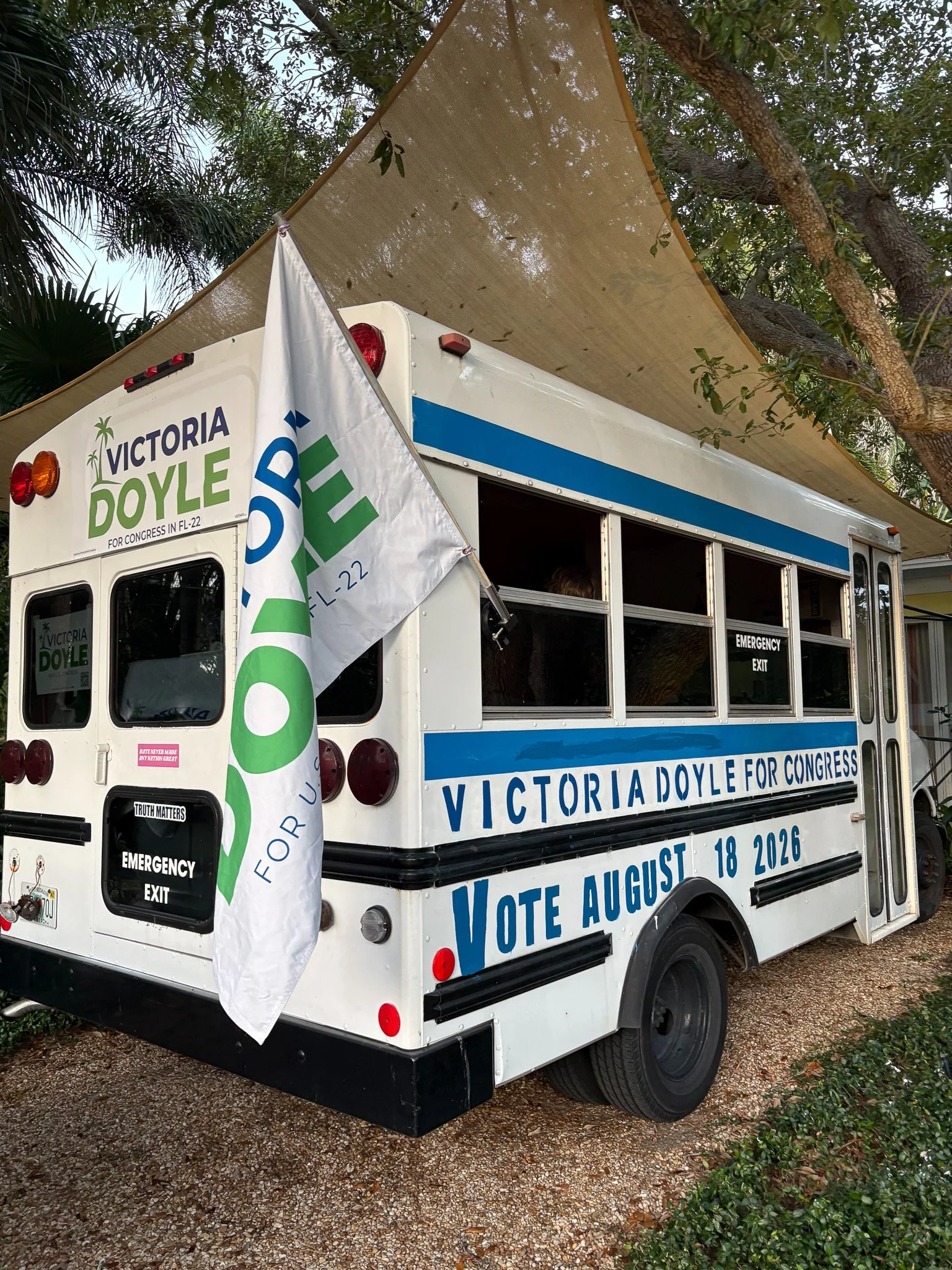 Mobile campaign vehicle with political signage for Victoria Doyle, advocating for voting on August 18, 2026, parked on gravel with a large tree and shade cloth overhead.