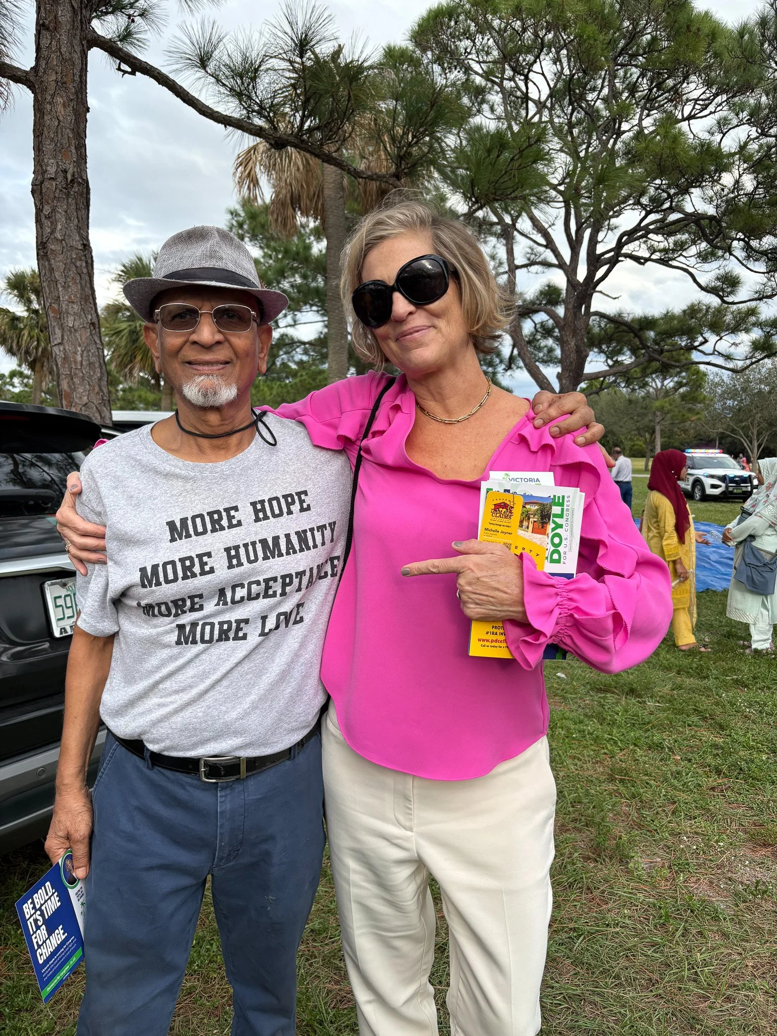 A man and woman standing outdoors among trees and other people, smiling. The man is wearing glasses, a gray hat, a gray T-shirt with black text, and dark pants. The woman is wearing large sunglasses, a pink blouse, and beige pants, holding campaign f